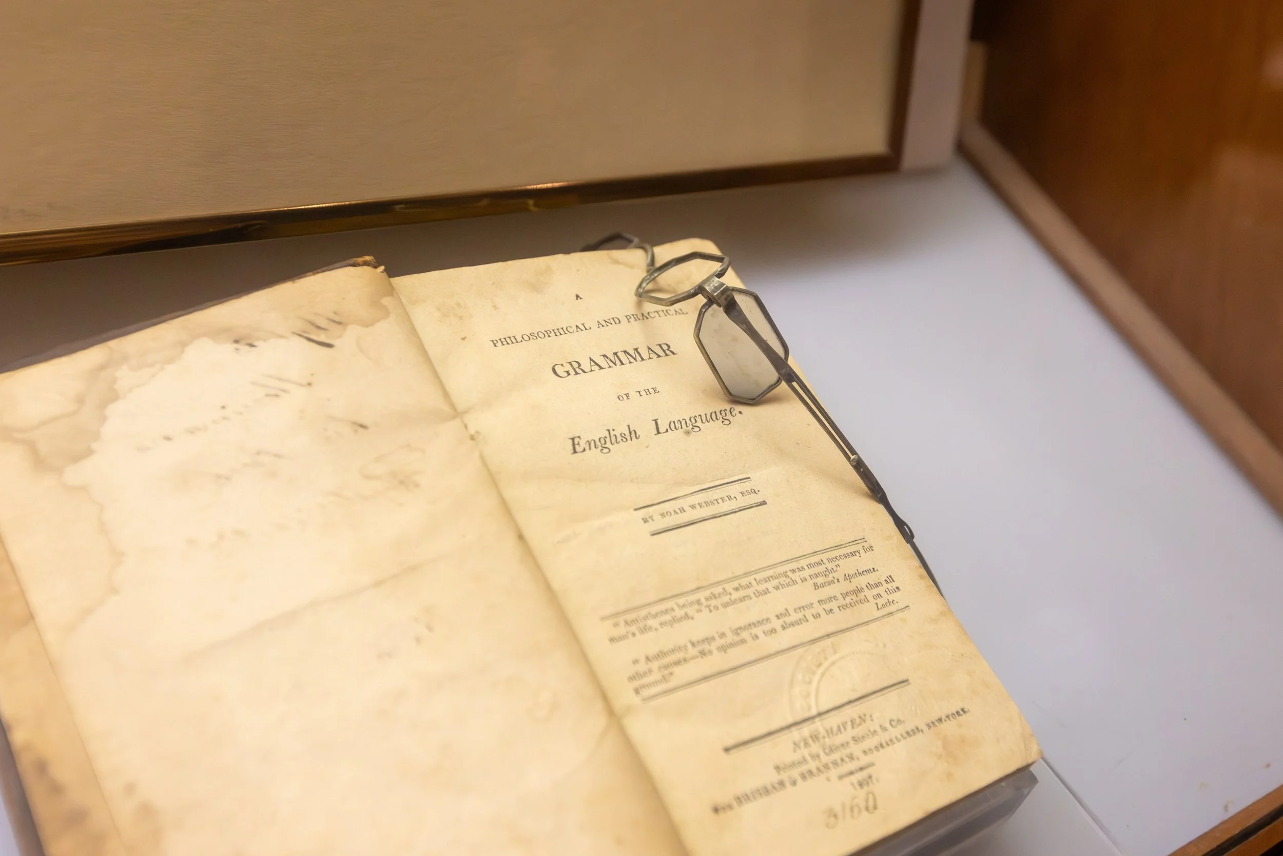 An old worn book titled 'Philosophical and Practical Grammar of the English Language' is open on a table, with a pair of metal eyeglasses resting on it.