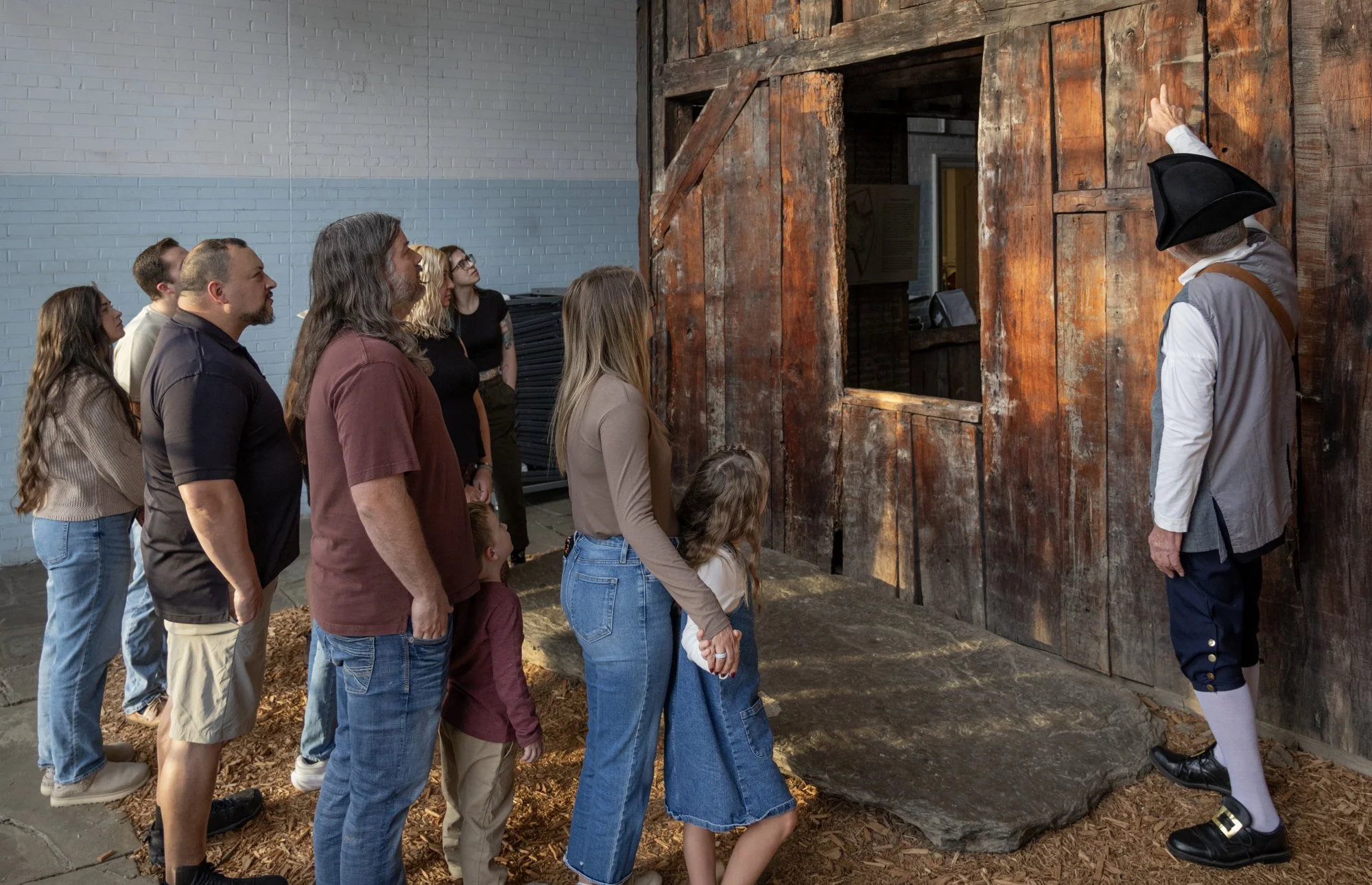 A man in colonial wear point to a wooden house while families listen to tour.