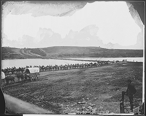 A black and white photo of a large group of horses and riders traveling along a road by a body of water, with a landscape of rolling hills in the background.
