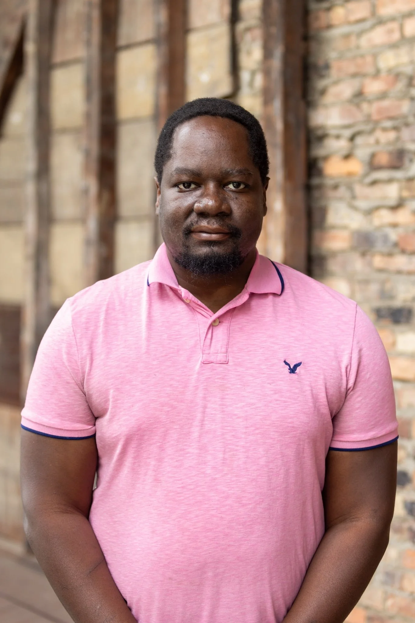A man with dark skin and short black hair wearing a pink polo shirt, standing in front of a brick and wooden wall.