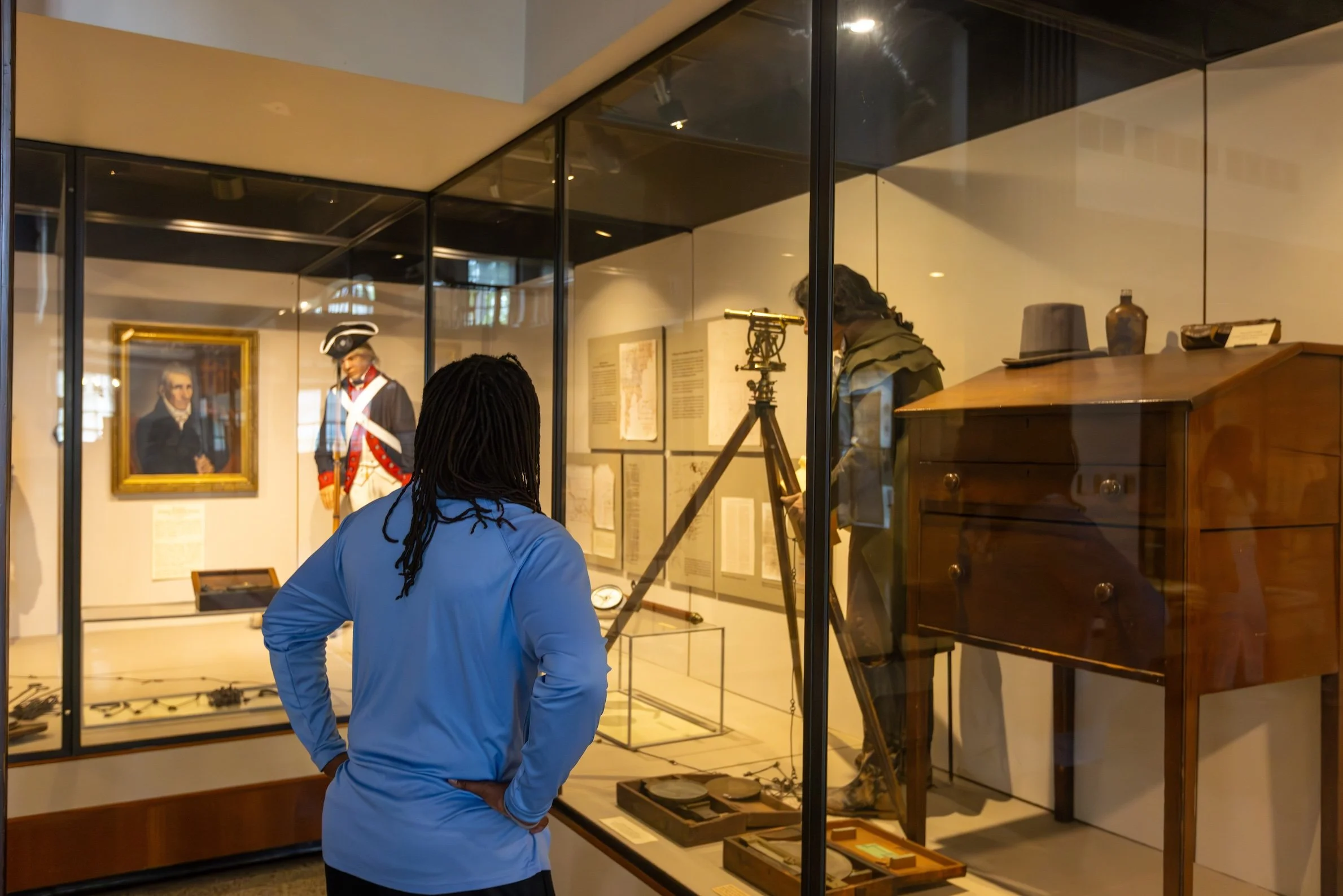 A person with long braids wearing a blue jacket is looking at historical artifacts displayed in a glass case at a museum. The artifacts include old maps, a mounted telescope, and other historical items.