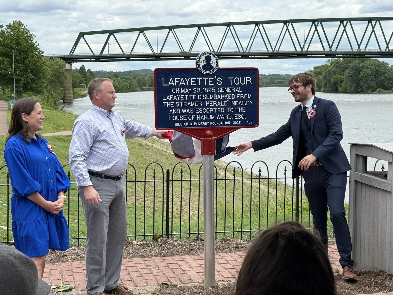 Three people standing beside a historical marker near a river, with a bridge above. The man on the right is unveiling the marker, and the woman on the left is watching, smiling. The man in the middle is pointing at the marker. The scene is outdoors, with greenery and cloudy skies in the background.