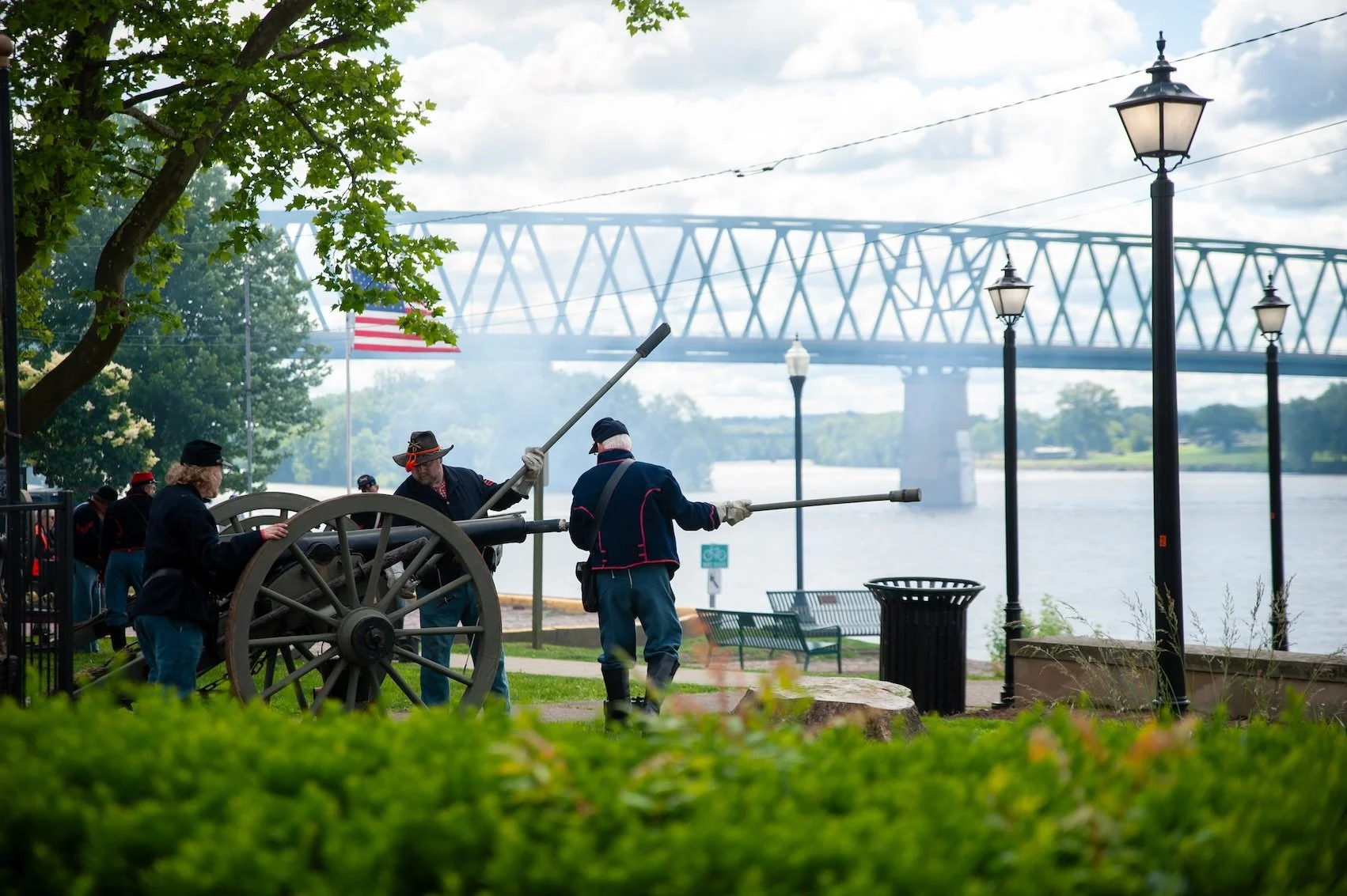 Historical reenactors dressed in Civil War uniforms operating a cannon near a river, with a bridge and street lamps in the background.