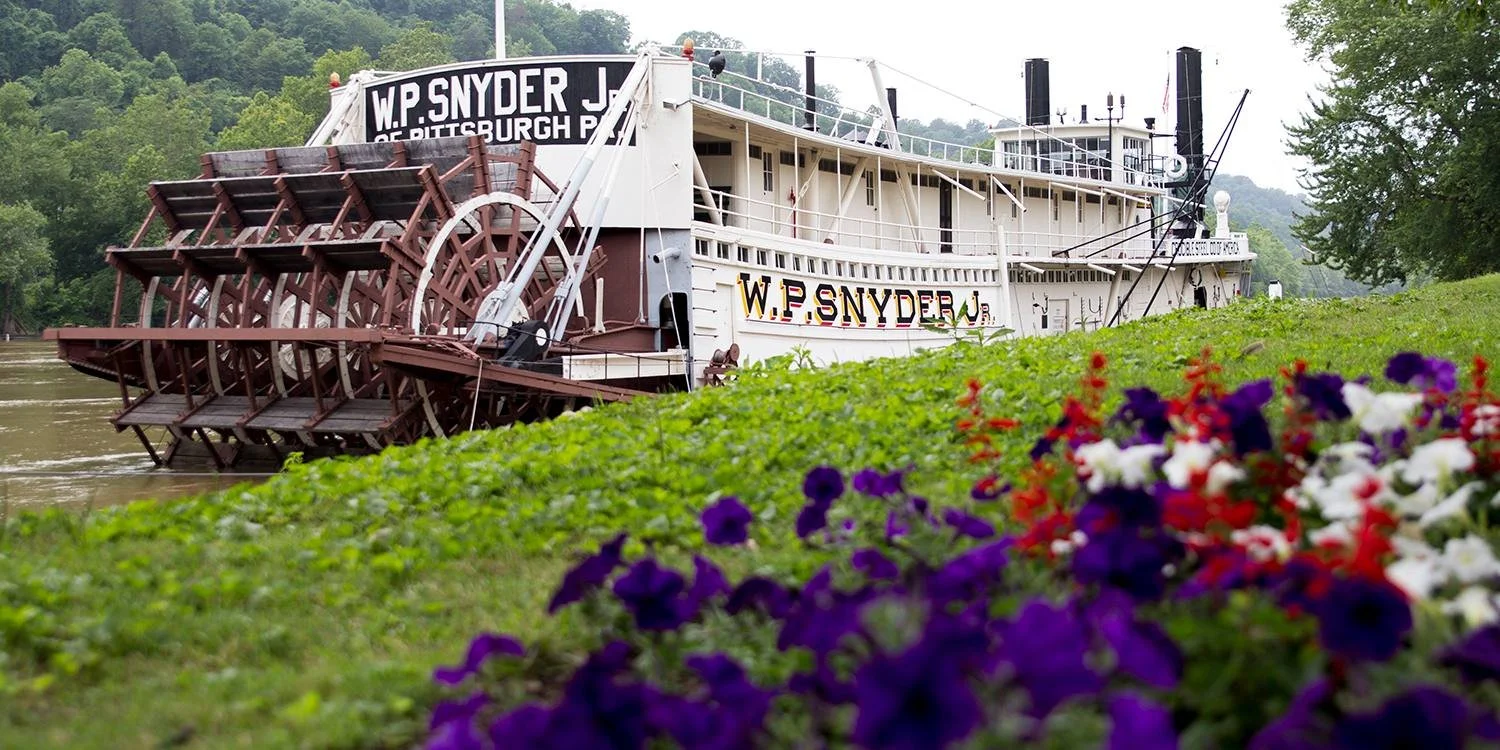 A historic paddlewheel riverboat named W.P. Snyder Jr. floating on a river with green hills in the background. The boat's name is displayed on the side and a black sign on top. In the foreground, there is a grassy area with colorful purple, red, white, and purple flowers.