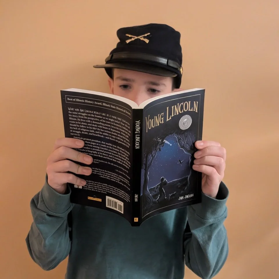 A young boy wearing a black cap with gold insignia and a gray hoodie is reading a book titled 'Young Lincoln' by Jan Jacob.