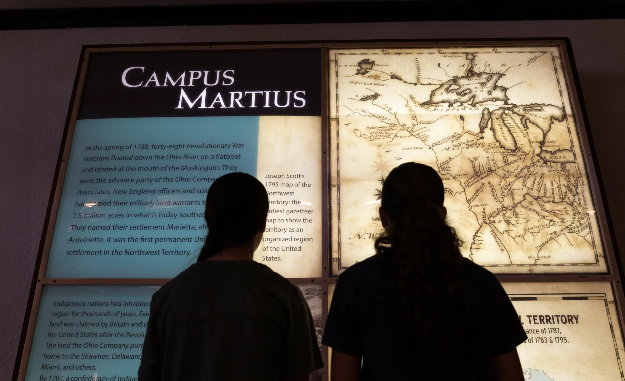 Two girls stand in in front of a lit museum panel. The panel gives historic facts and displays maps.
