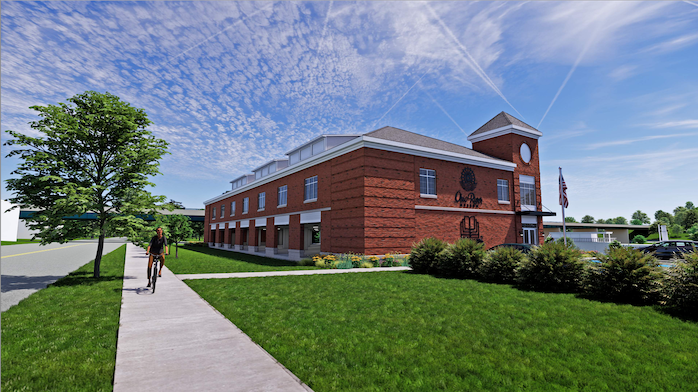 A large red-brick building with a clock tower on the right and an American flag flying, surrounded by green grass and trees, with a sidewalk and a person walking on it under a partly cloudy blue sky.