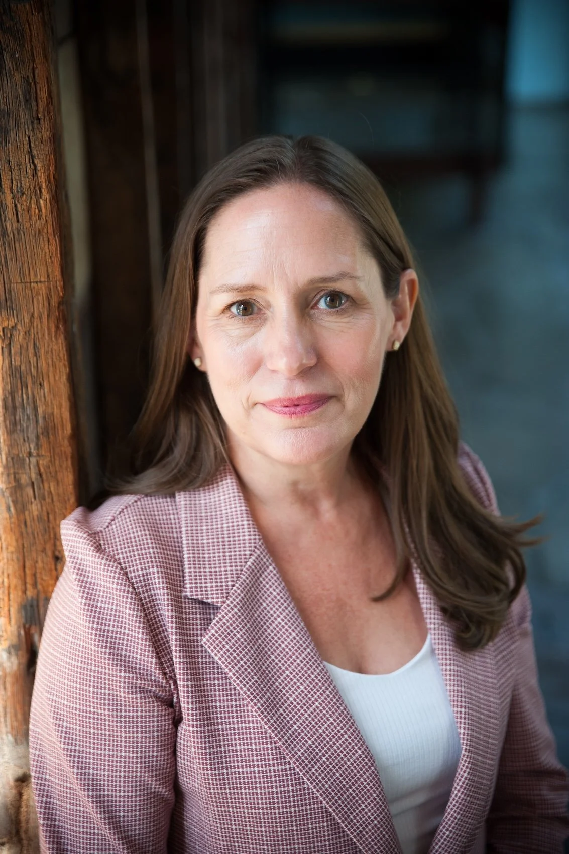 A woman with shoulder-length brown hair, wearing a pink checkered blazer and white top, sitting next to a wooden post, looking directly at the camera.