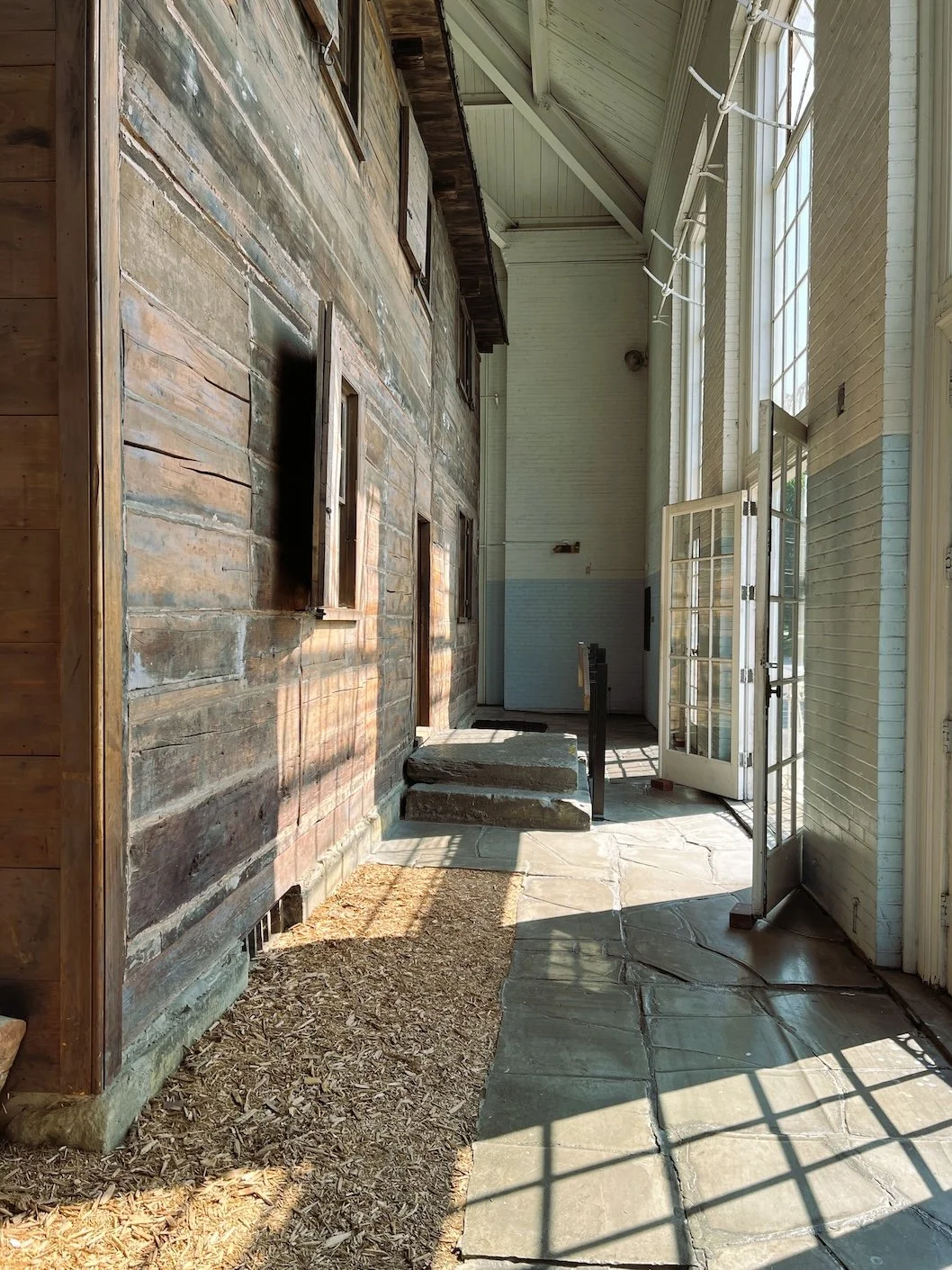 Sunlit enclosed porch with brick and wooden walls, open French doors, stone flooring, and steps leading to a door inside.