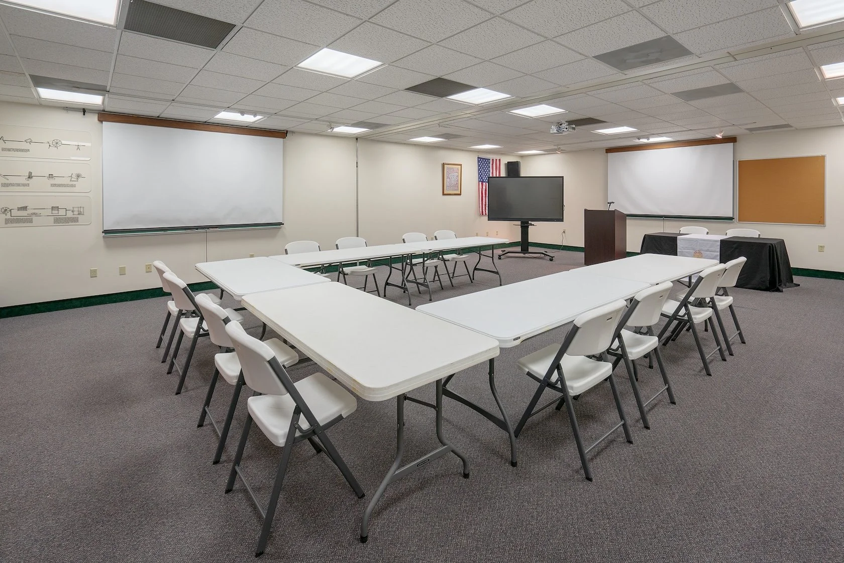 Empty conference room with white tables arranged in a U-shape, white chairs, a large screen on a stand, whiteboards, and American flags on the wall.