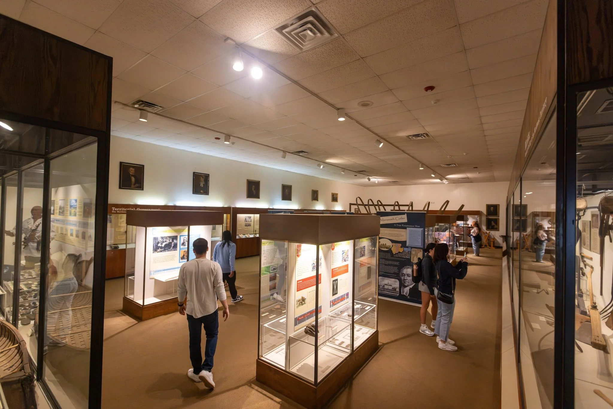 People exploring exhibits in a museum with display cases and informational panels, framed portraits hanging on the walls, and museum artifacts.