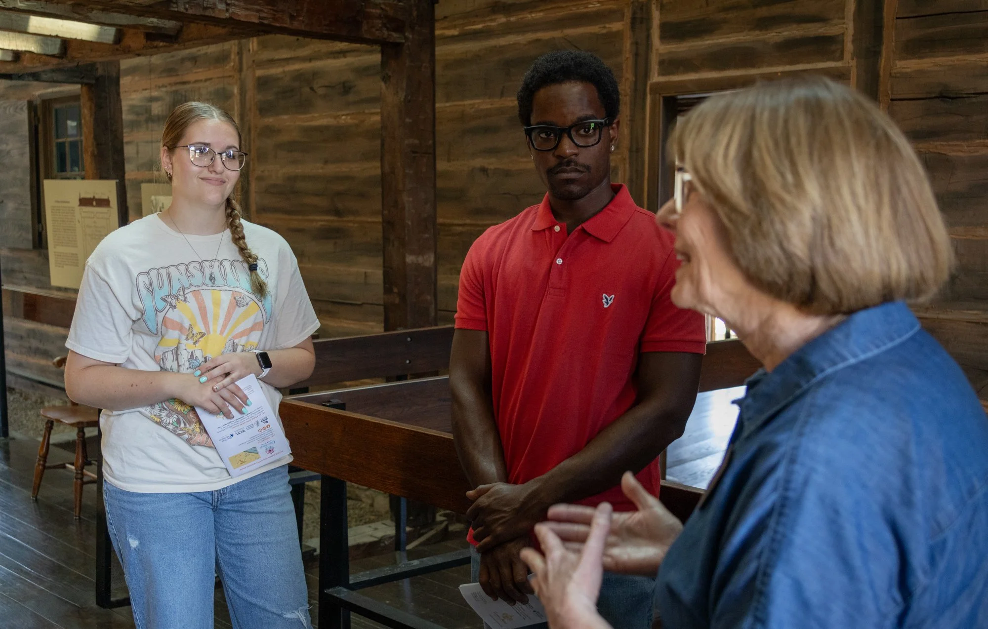 An older woman in a blue top speaks to an African American man with black glasses and a red shirt and a young Caucasian female with glasses, a white shirt and blue jeans.