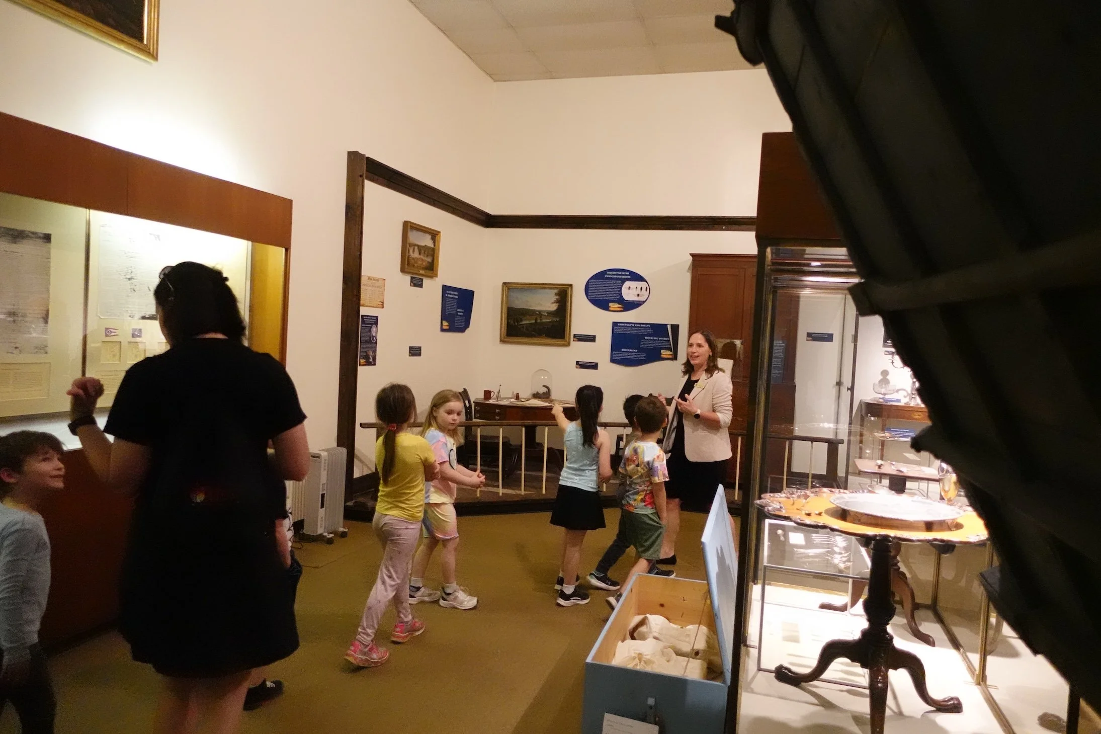 A museum exhibit room with a woman talking to children, some standing and some sitting, surrounded by historical display artifacts and informational panels.