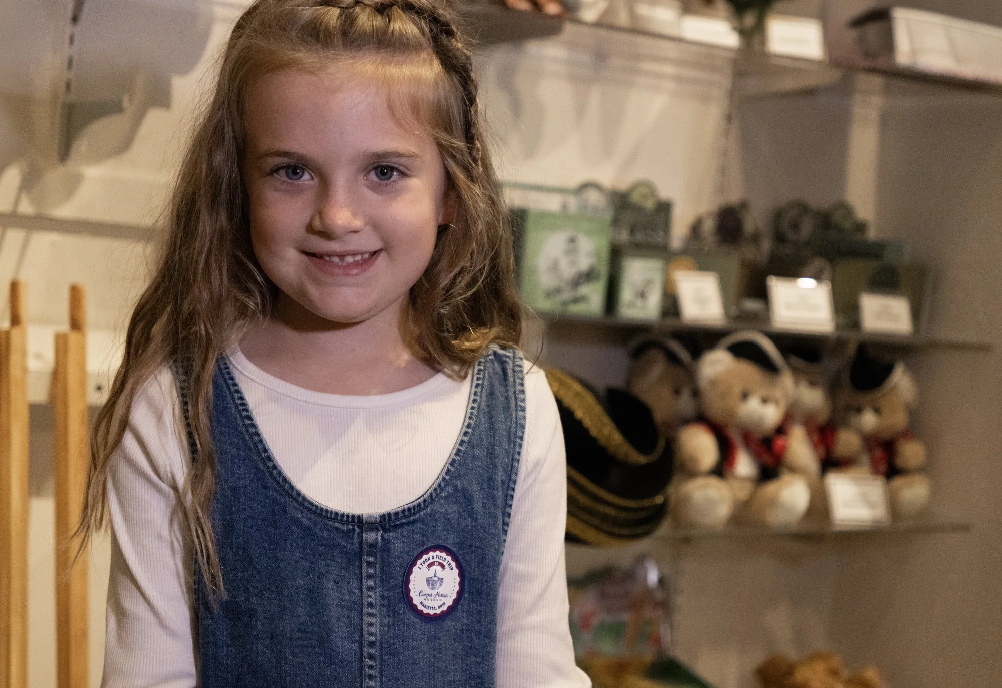 A young girl stands in a gift shop with stuffed animals and hats. She has long brown hair and wears a white top, a blue jean dress, and a sticker.