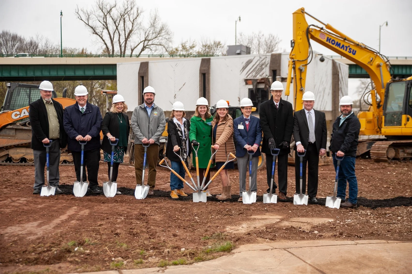 Group of people wearing hard hats standing with shovels at a groundbreaking ceremony on a construction site with heavy machinery in the background.