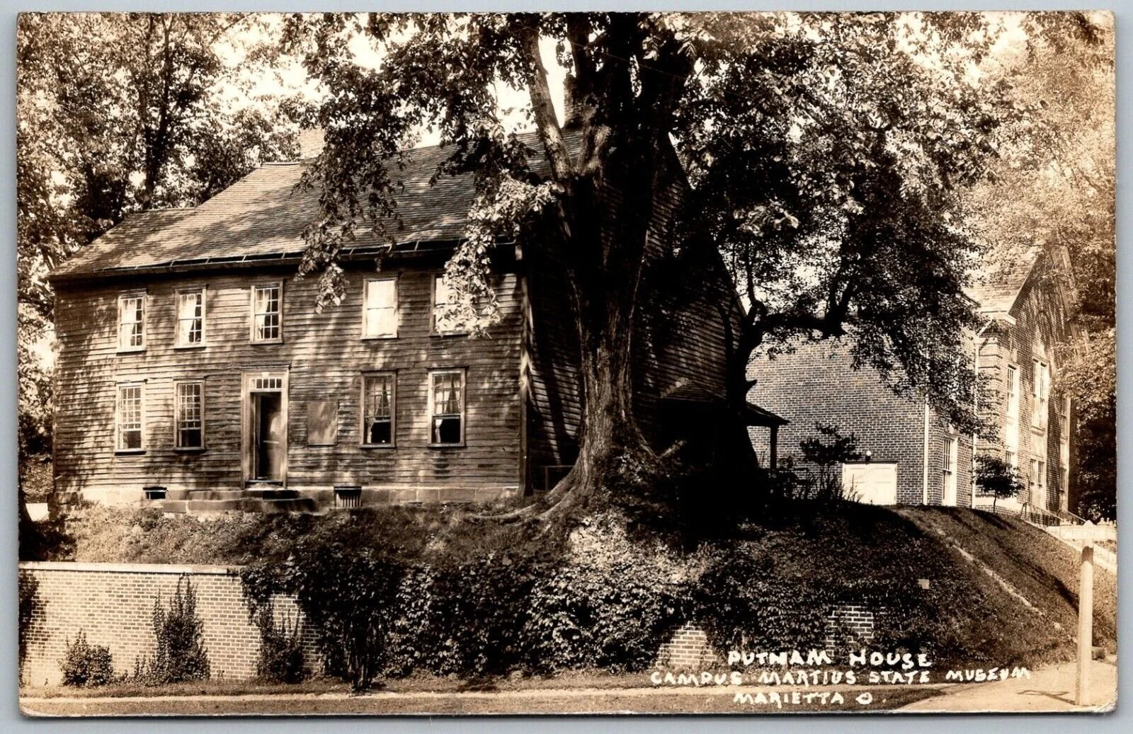 A sepia photograph of a large, old wooden house with multiple windows, partly covered by a big tree in the yard. A brick building is visible behind the house. The grass around the house is slightly overgrown, and the house is raised on a small hill with a brick retaining wall at the front. Handwritten text at the bottom right corner
