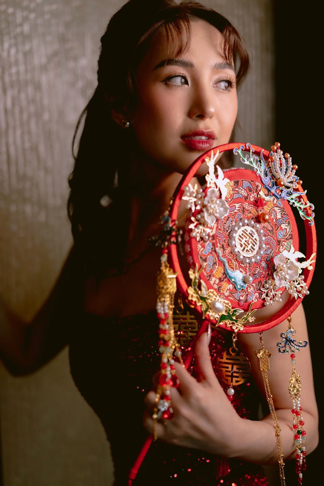 A woman with curly brown hair and red lipstick is holding a decorative circular item with Chinese characters, pearls, and colorful ornaments, wearing a black dress with red sequins.