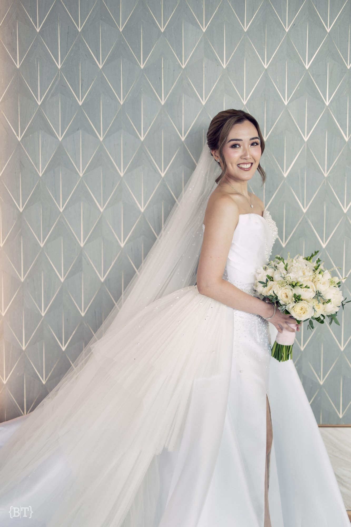 Bride in a white wedding dress holding a bouquet of white roses, smiling, with a geometric-patterned wall in the background.