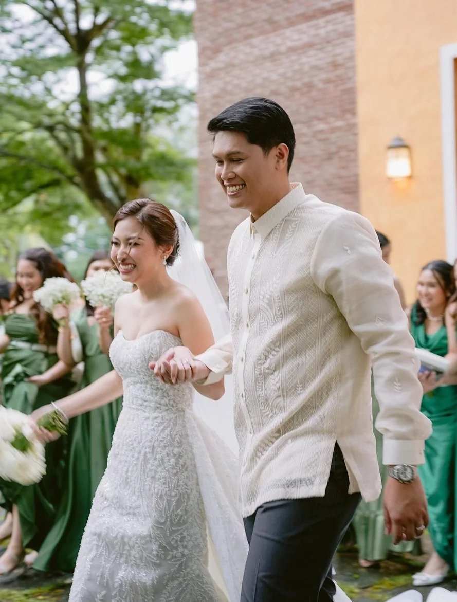 A happy bride and groom holding hands and walking outdoors during their wedding celebration, surrounded by friends in green dresses.