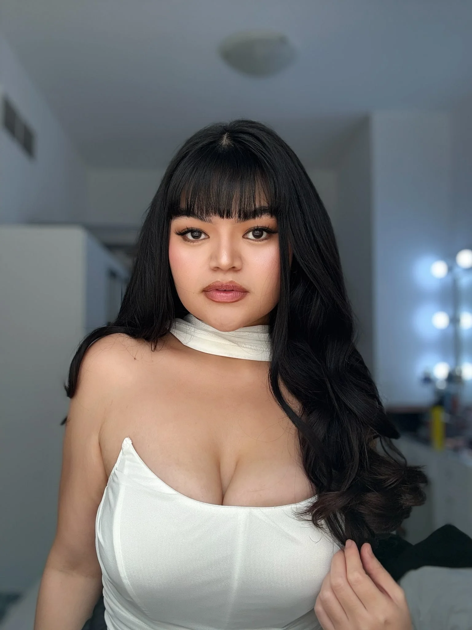 A young woman with long black hair and bangs, wearing a white strapless top and a matching white scarf around her neck, in a room with light-colored walls and background lighting.