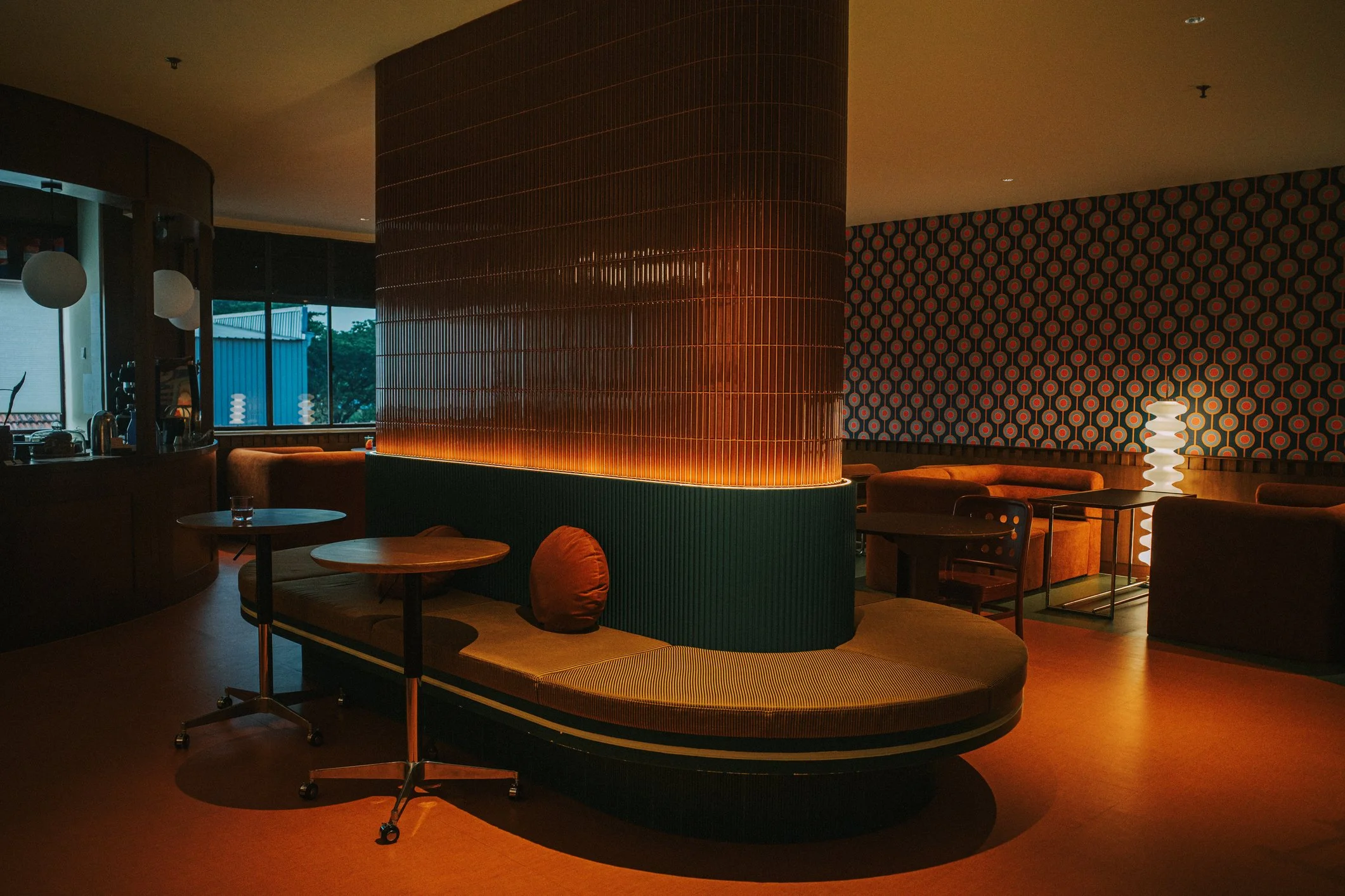 A lounge area with curved seating, small round tables, and a decorative divider with orange lighting. The background includes patterned wallpaper and a unique white floor lamp, with a window showing an outdoor scene.