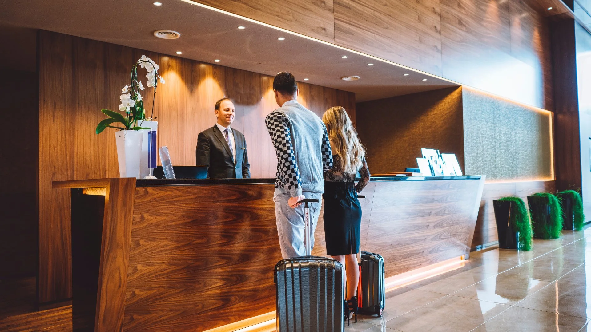 A man and woman with suitcases checking in at a hotel reception desk, with a reception staff member behind the counter.