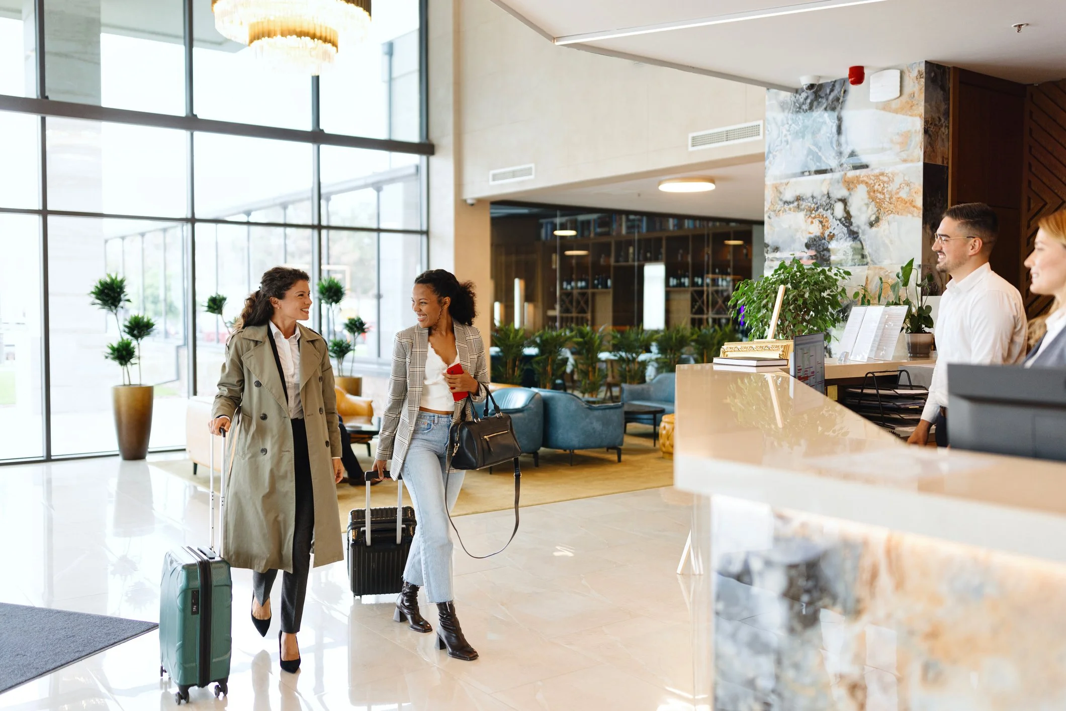 Two women with suitcases check in at a hotel reception desk while a male and female staff member assist them in a modern hotel lobby.