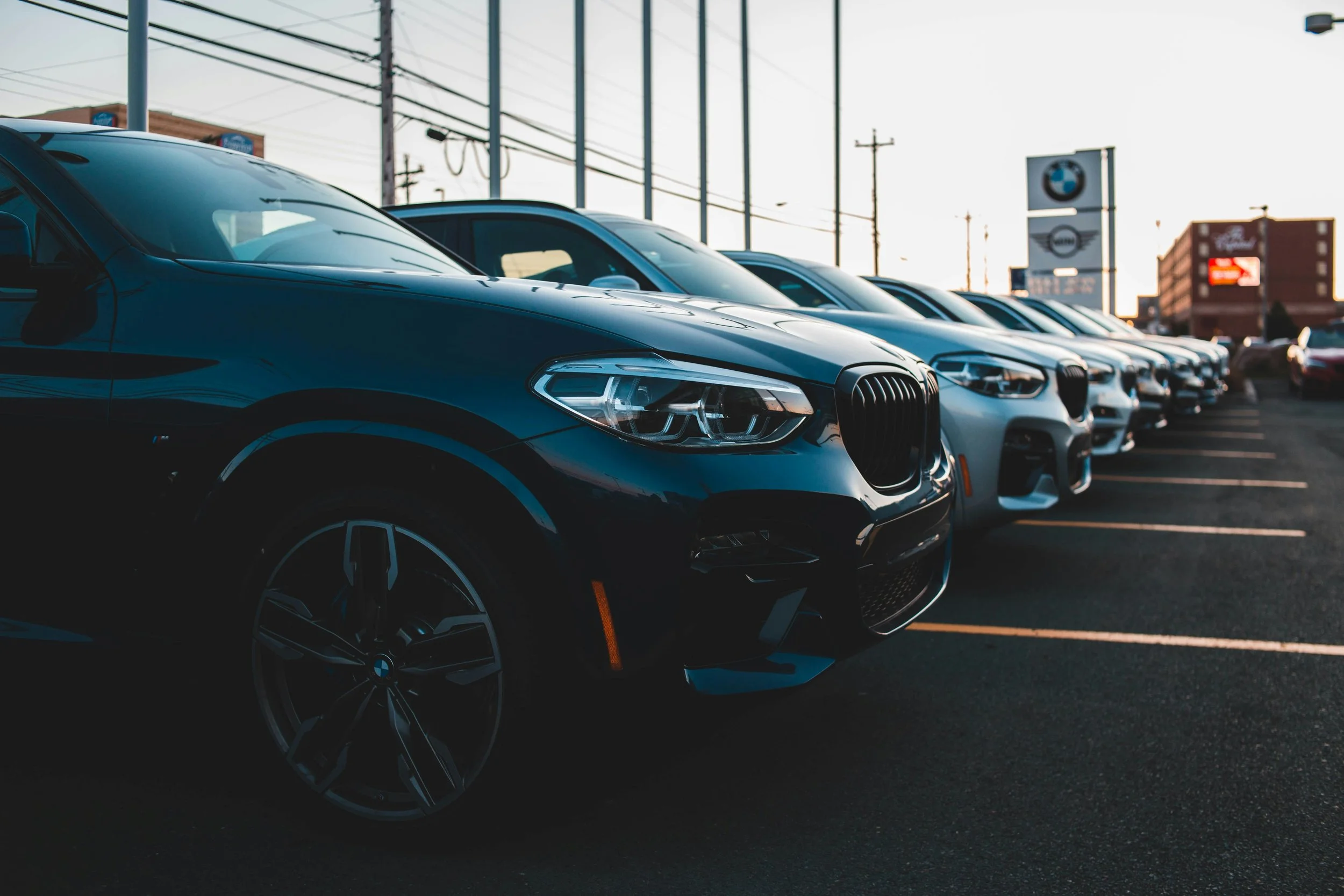 A row of black and silver cars parked outside a car dealership with BMW and MINI signs in the background during sunset.