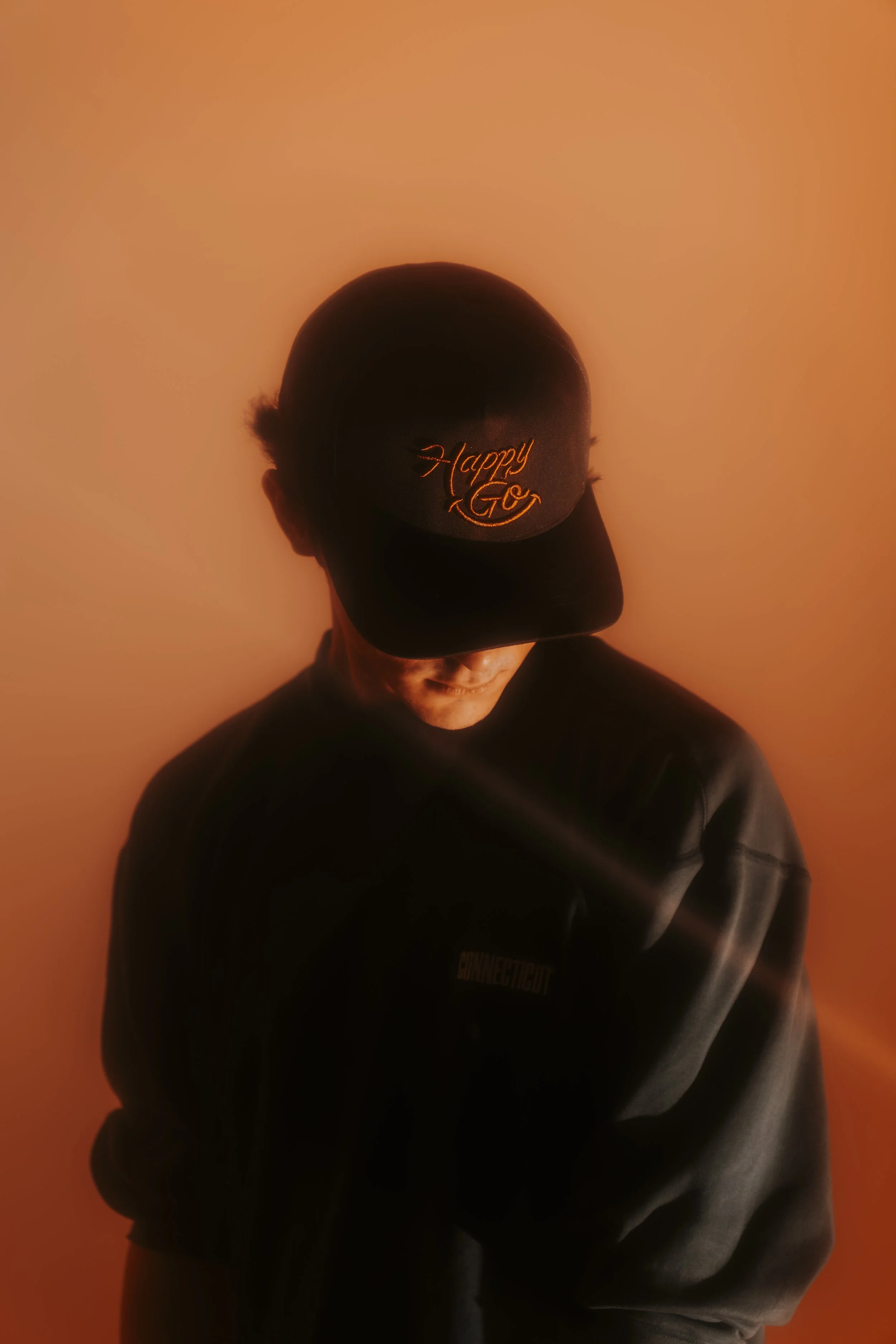 A person wearing a black cap with orange embroidered text and a black jacket, standing against a warm orange background, with their head tilted downward.
