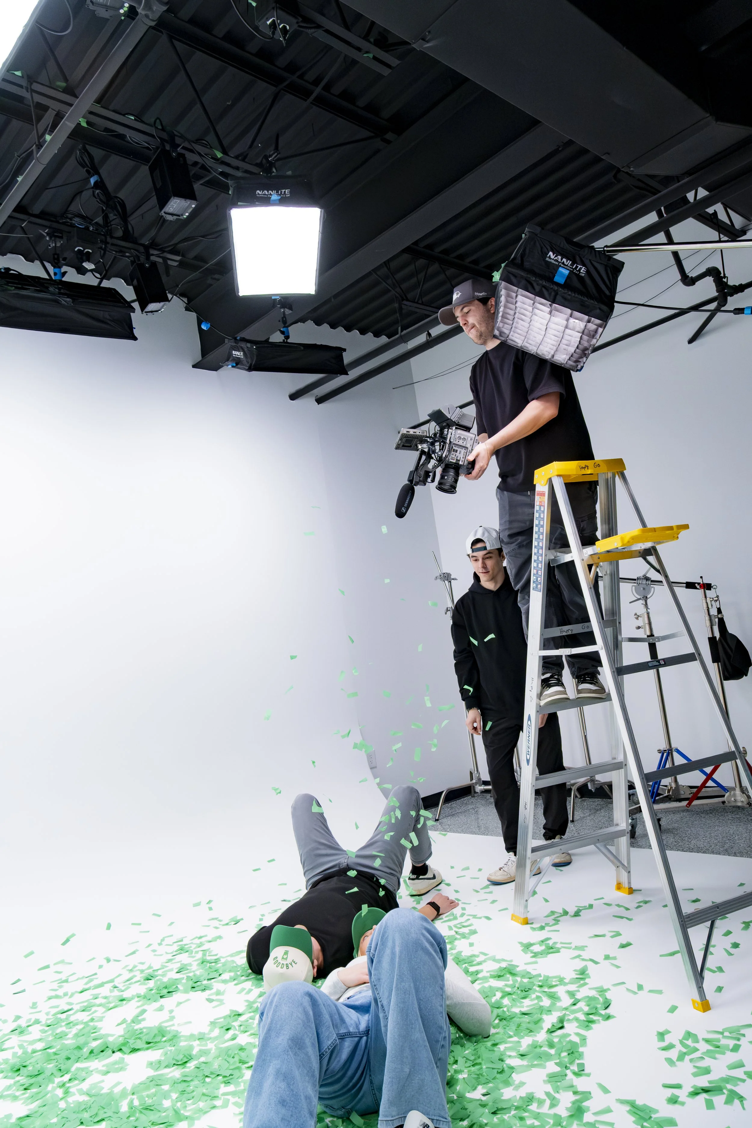 A video production set with two people standing on a ladder, filming a scene where two individuals are lying on the floor amidst green confetti. Studio lighting equipment is overhead, and a plain white backdrop is in the background.