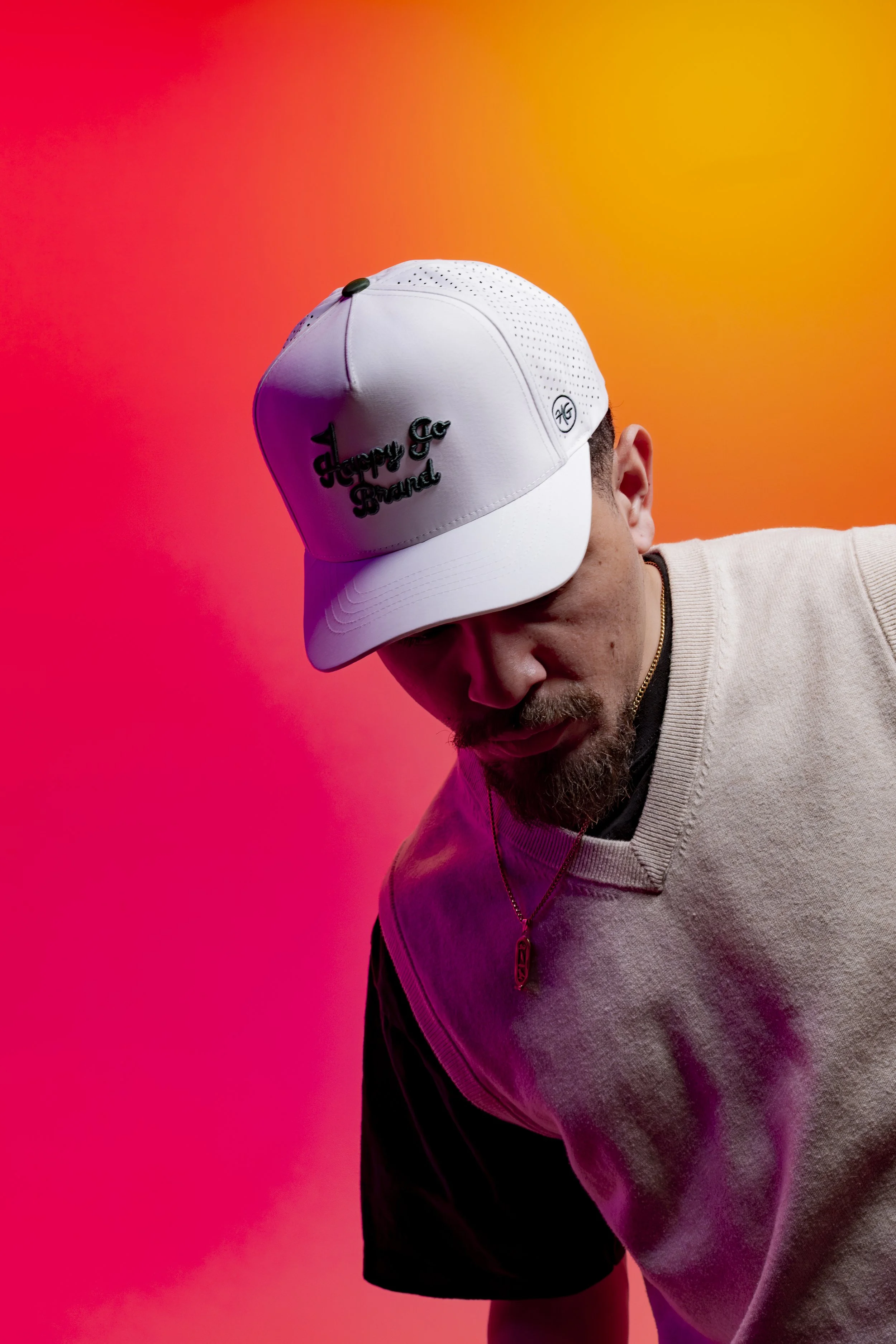 Young man standing in front of a colorful background with a white Happy Go Brand golf hat on. 