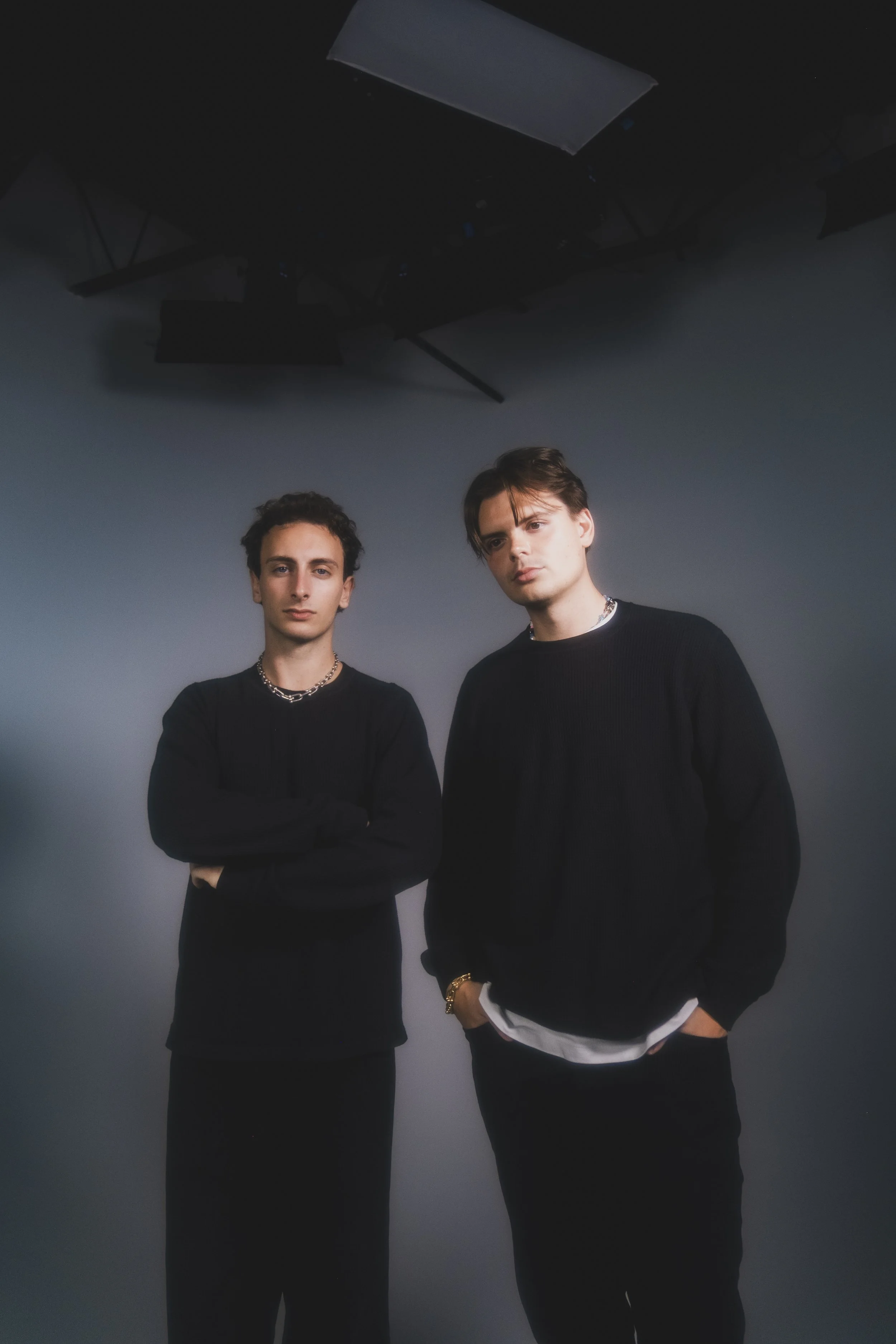Two young men standing in a photo studio with gray background, both wearing black sweaters, one with arms crossed and the other with hands in pockets, studio lighting overhead.