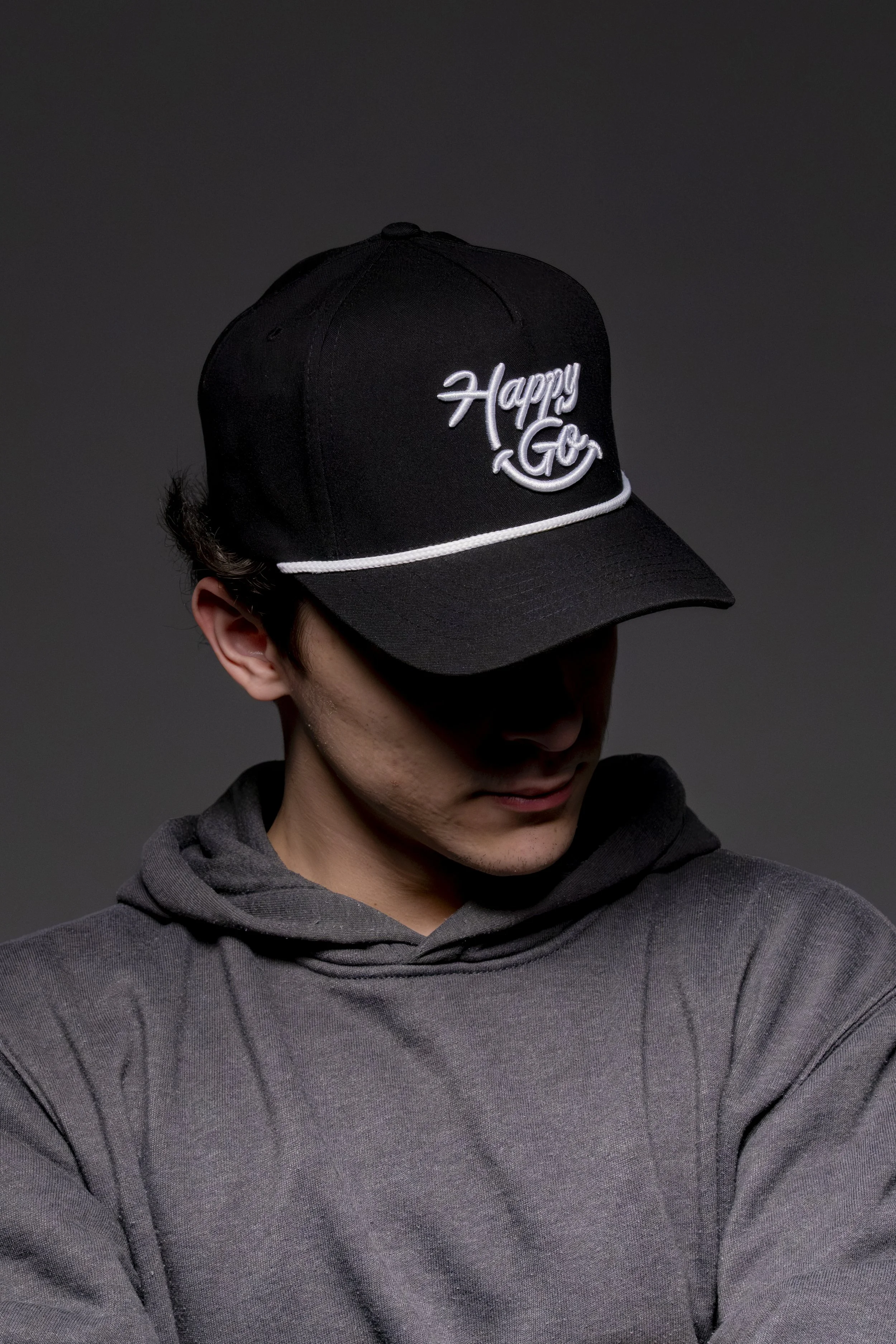 A young man wearing a black cap with white embroidered text that reads 'Happy Go' and a dark gray hoodie, looking downward against a dark background.
