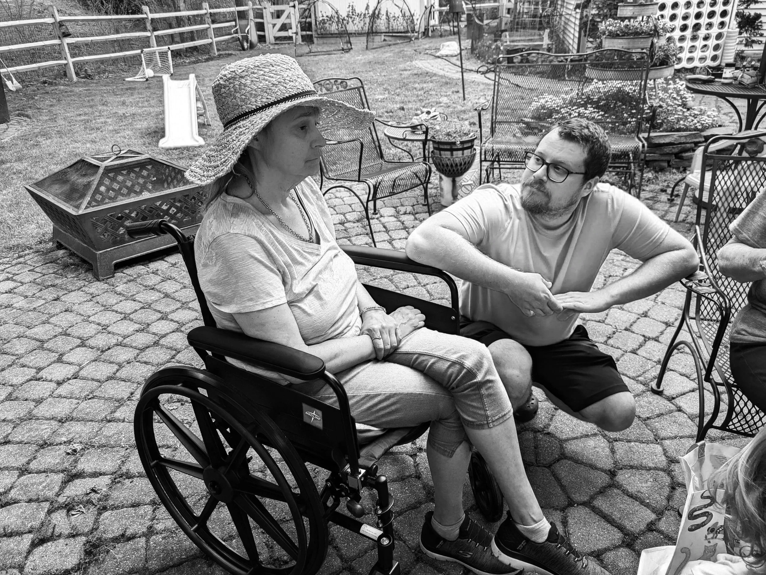 A woman in a wheelchair wearing a sun hat talks with a man with glasses sitting nearby in a garden patio with outdoor furniture.