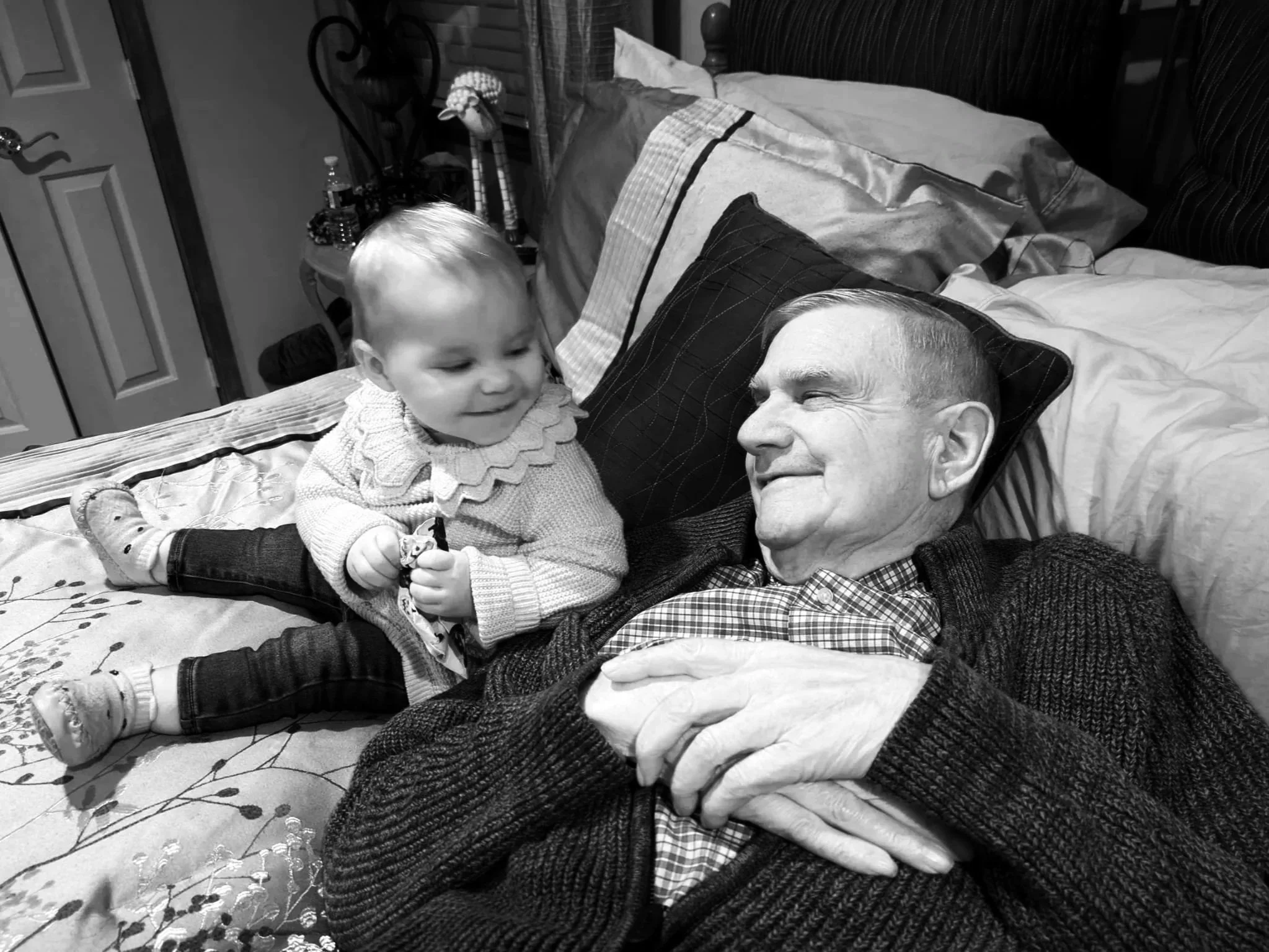 A young child with light-colored hair lying on a bed, turned towards an older man with short hair, smiling as they look at each other. The bed has multiple pillows and a blanket with a floral pattern, and a stuffed toy is visible near the child's feet. The room shows a nightstand with water bottles and other items, a closed door, and some decorative objects.