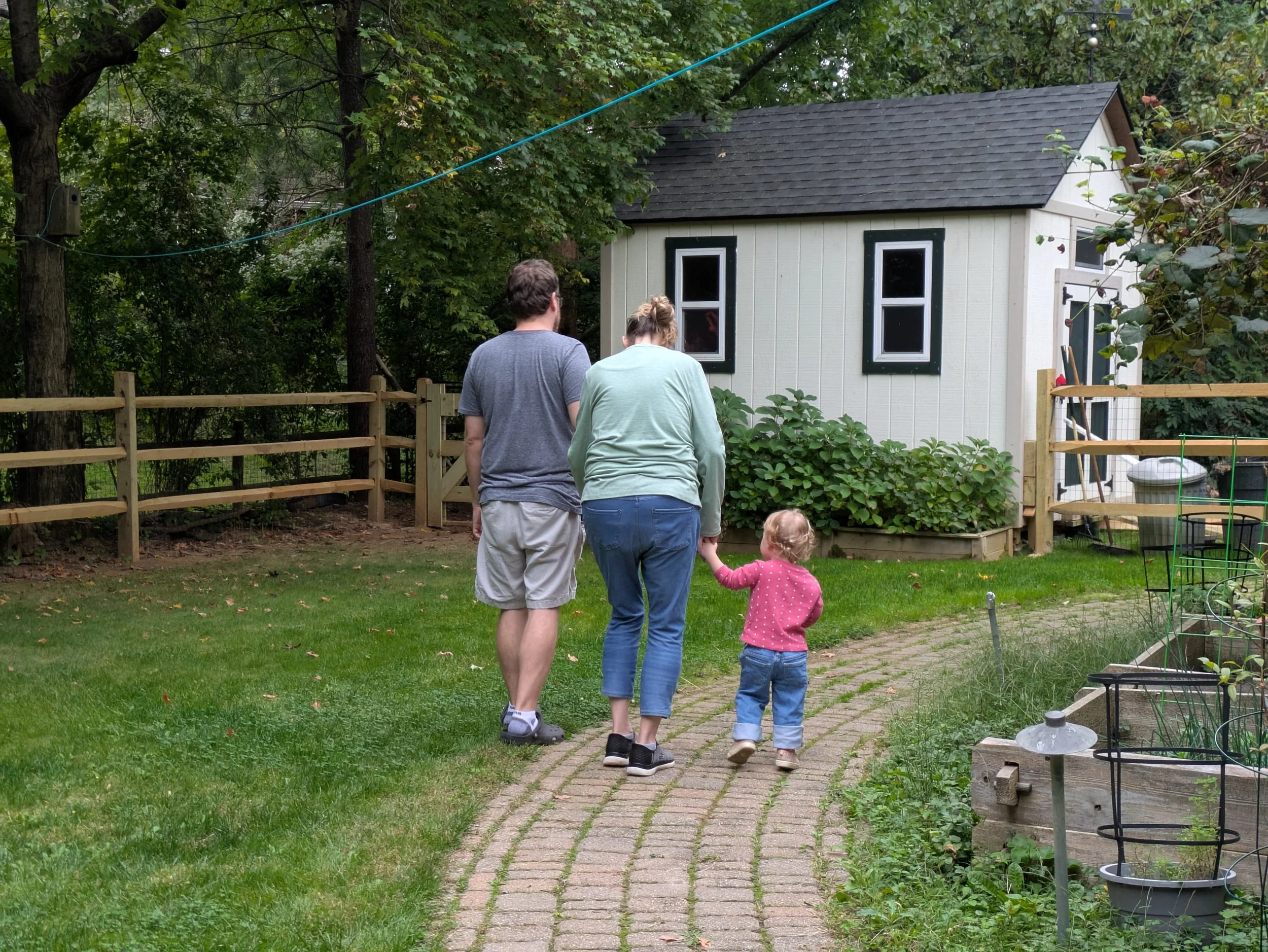 Three adults and two children walk down a brick pathway in a backyard with a small white shed and trees. The adults are viewed from behind, and the children are holding hands with one of the adults.