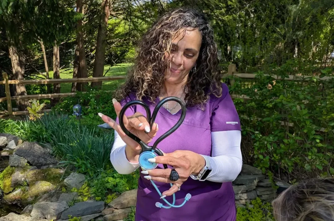 Woman with curly hair wearing purple scrubs holds a stethoscope with the heart-shaped tubing in an outdoor garden setting.