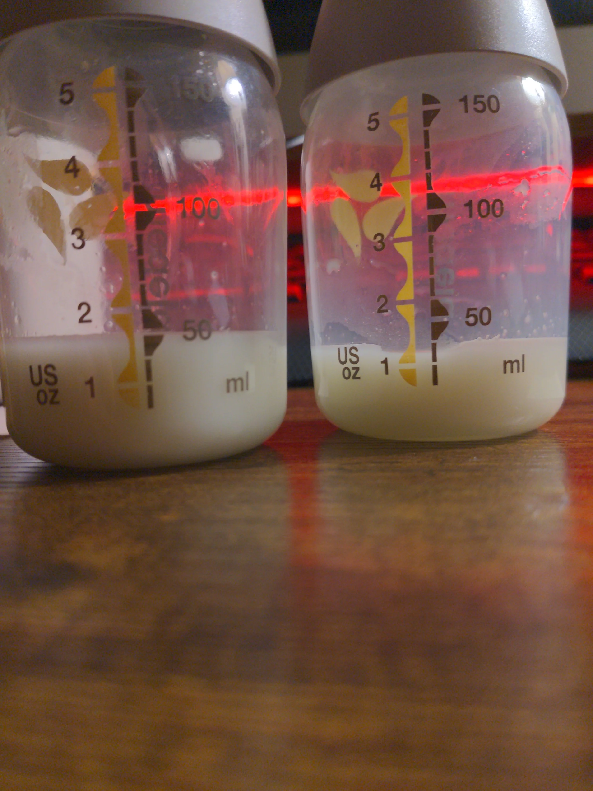 Two baby bottles filled with milk placed side by side on a wooden surface with measurement markings in milliliters and ounces, illuminated by a red light.