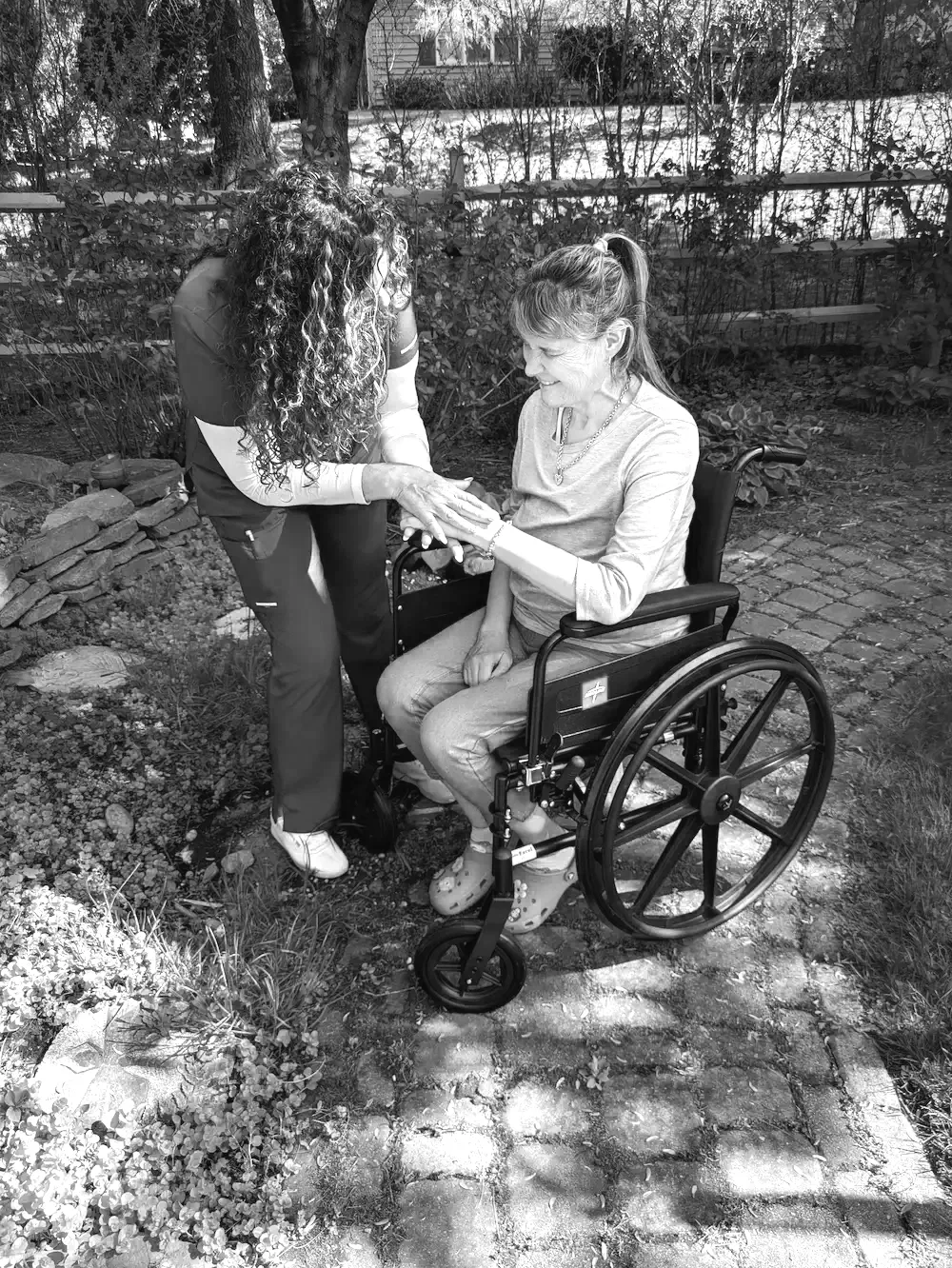 A woman in a wheelchair sharing a joyful moment with a caregiver outdoors in a garden. The caregiver is holding the woman's hand and leaning in.