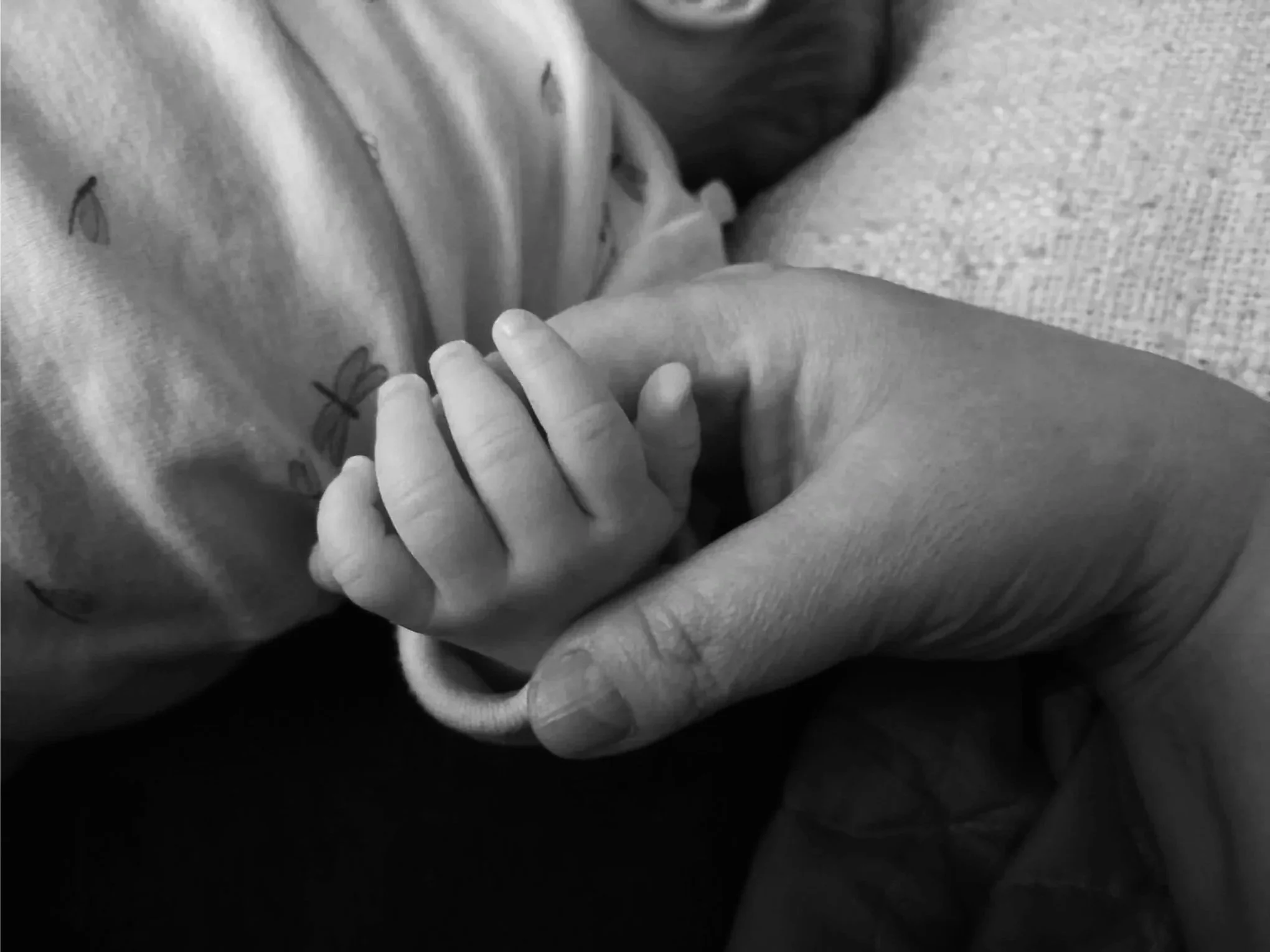Close-up black and white photo of a person gently holding a baby's hand, with the baby's face partially visible in the background.