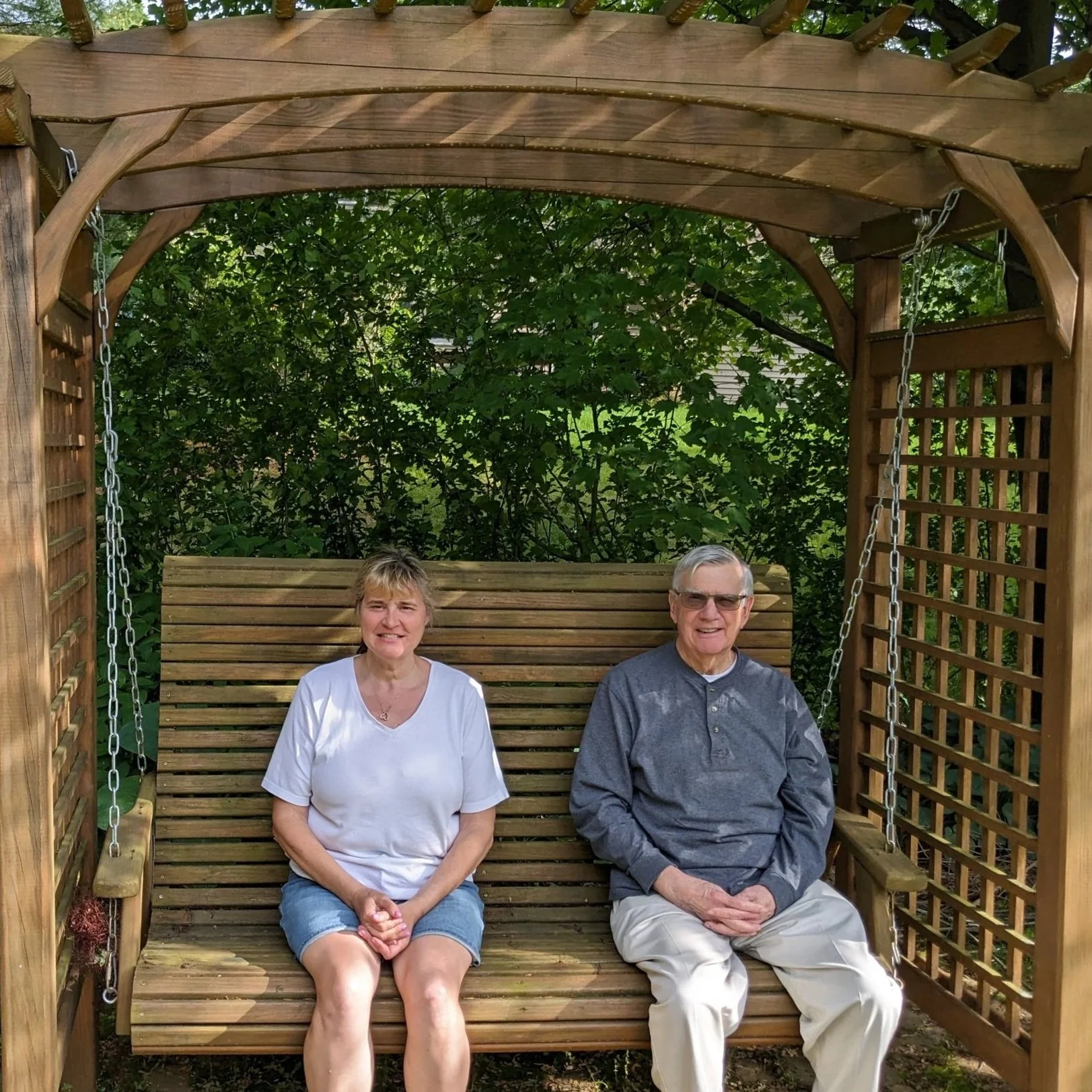An older woman and man sitting on a wooden swing with a wooden frame and chains in a garden with green foliage in the background.