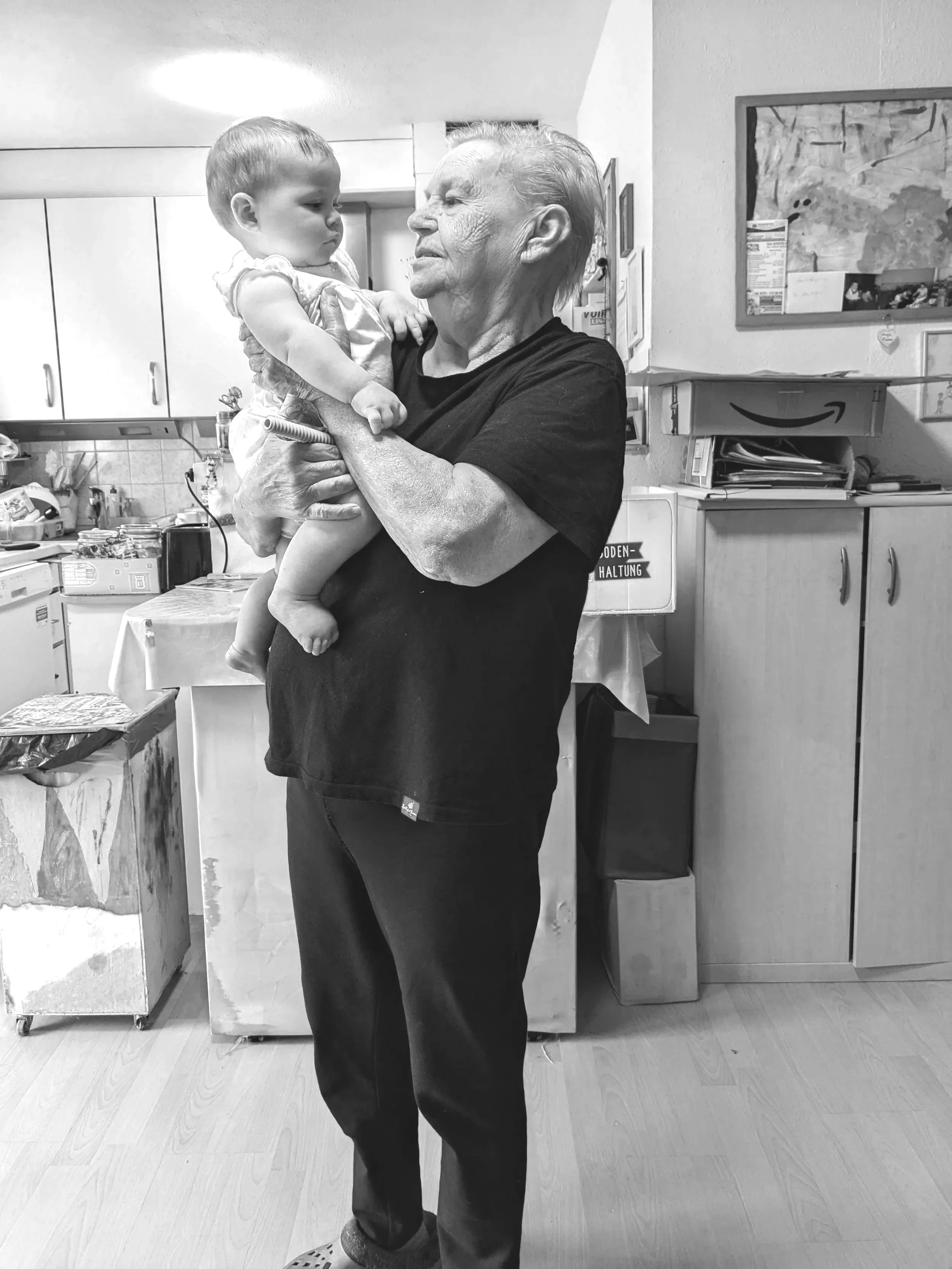 An elderly woman holding a young child in a kitchen, both appearing to gaze at each other softly.