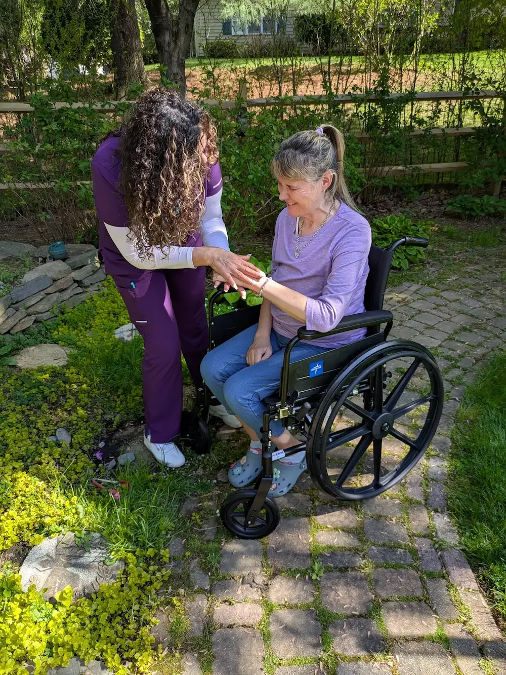 A caregiver and an elderly woman in a wheelchair sharing a joyful moment outdoors in a garden.