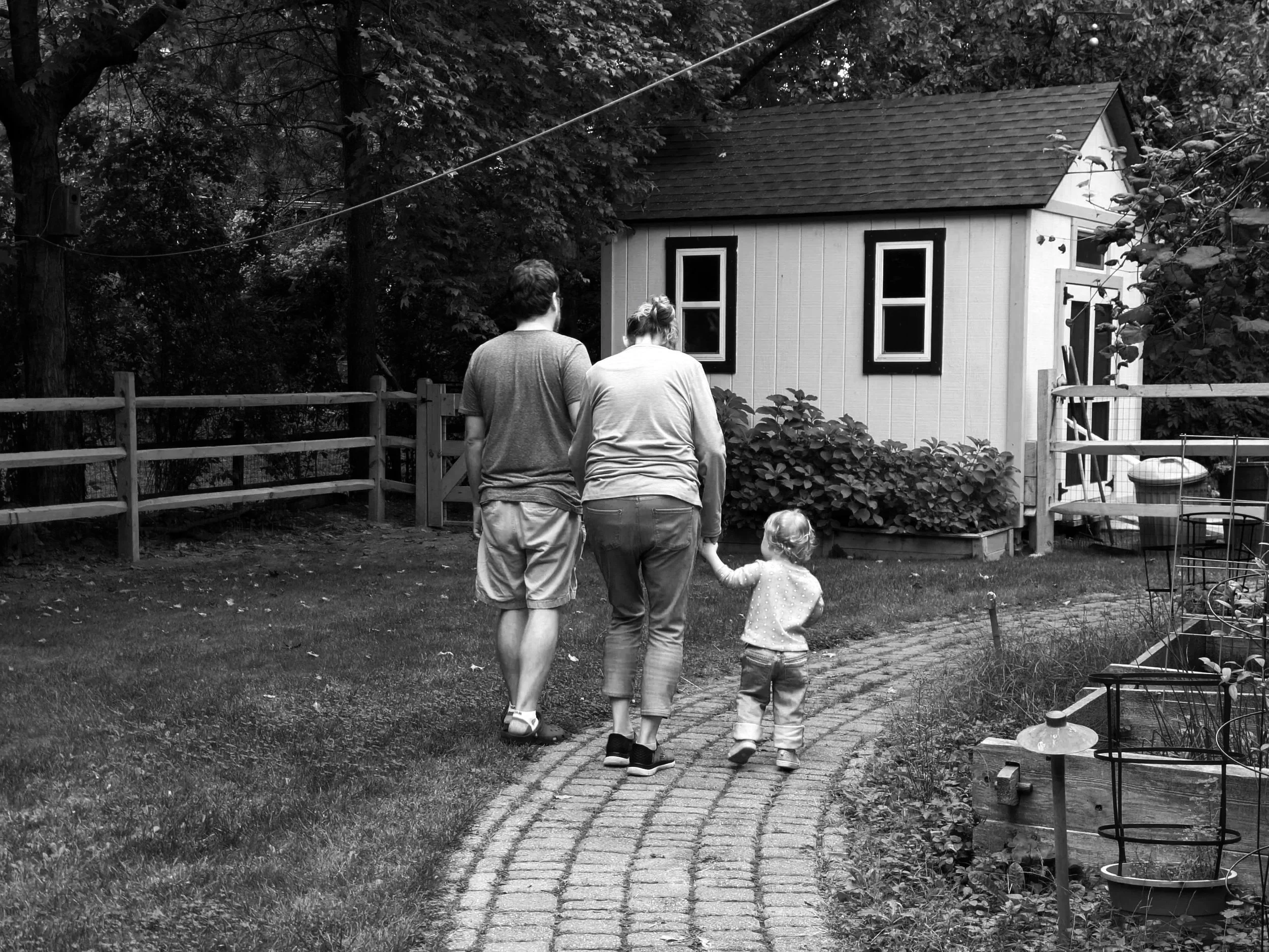 A black and white photo of two adults and three children walking along a brick path in a backyard, with a small house, trees, bushes, and garden furniture around them.