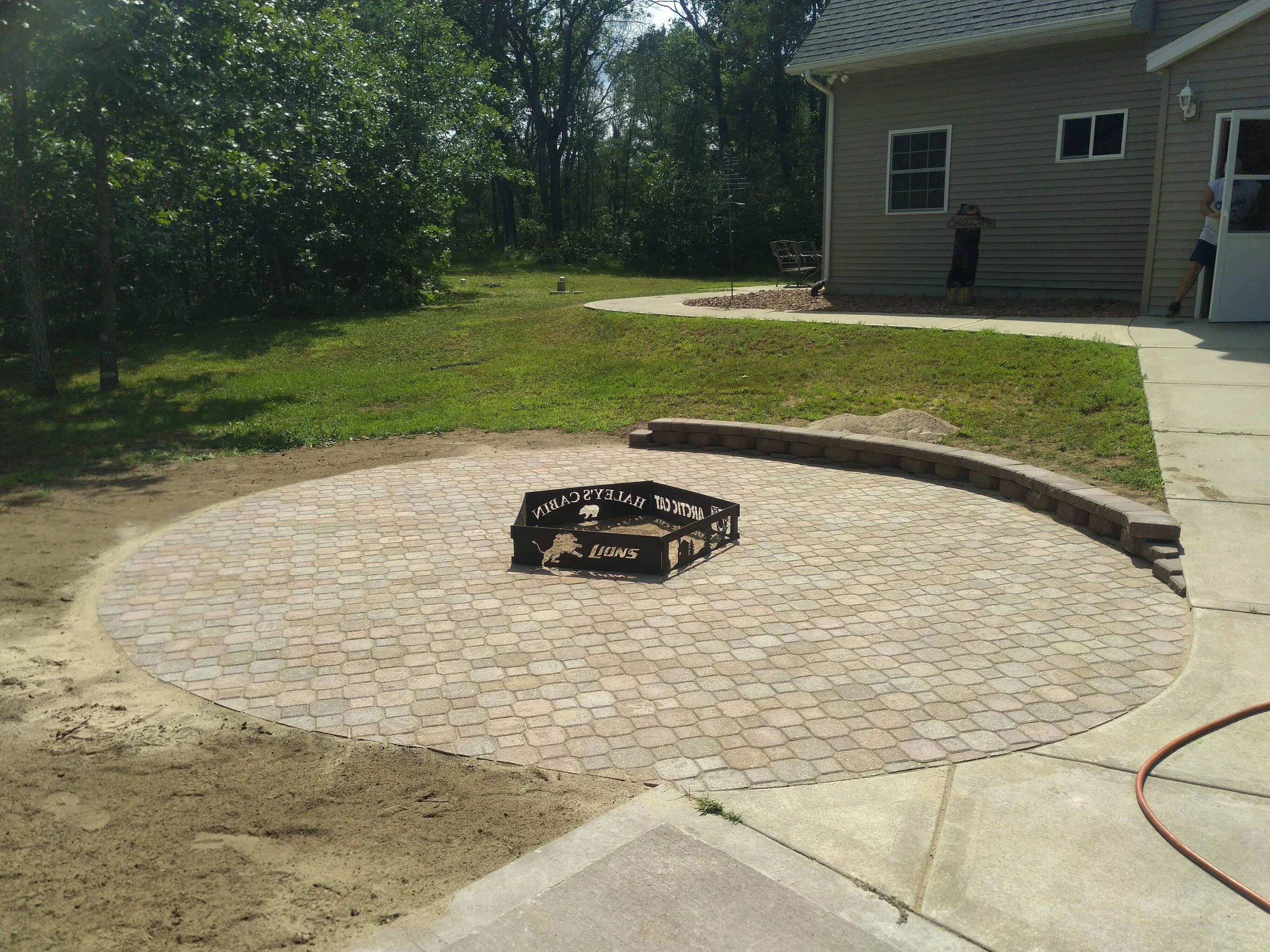A backyard patio with a circular brick paver section, a fire pit with a Michigan Wolverines logo, a brick retaining wall, a grassy lawn, a house with a sidewalk, and trees in the background.