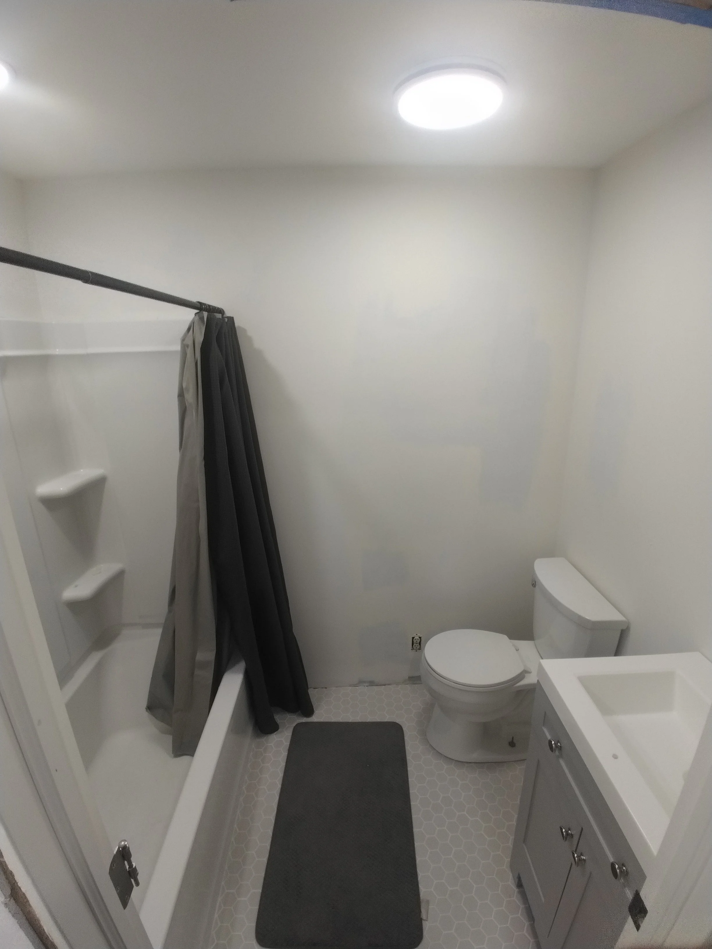 Small bathroom with a shower stall, toilet, gray rug, and a white vanity sink. Ceiling light fixture and plain white walls.