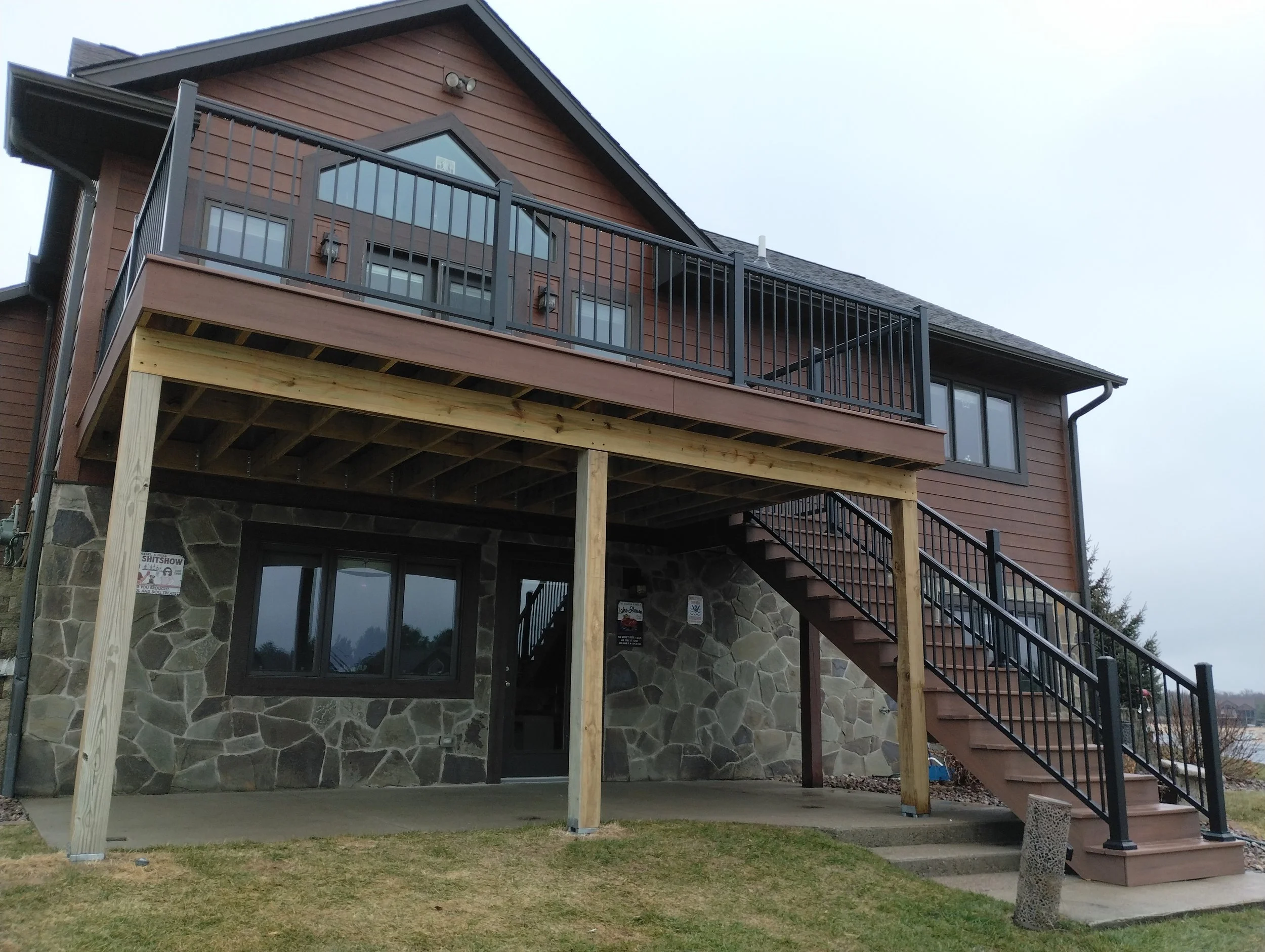 Multi-story house with a stone foundation, wooden siding, a large wooden deck with black metal railings, and an outdoor staircase leading to the ground level.