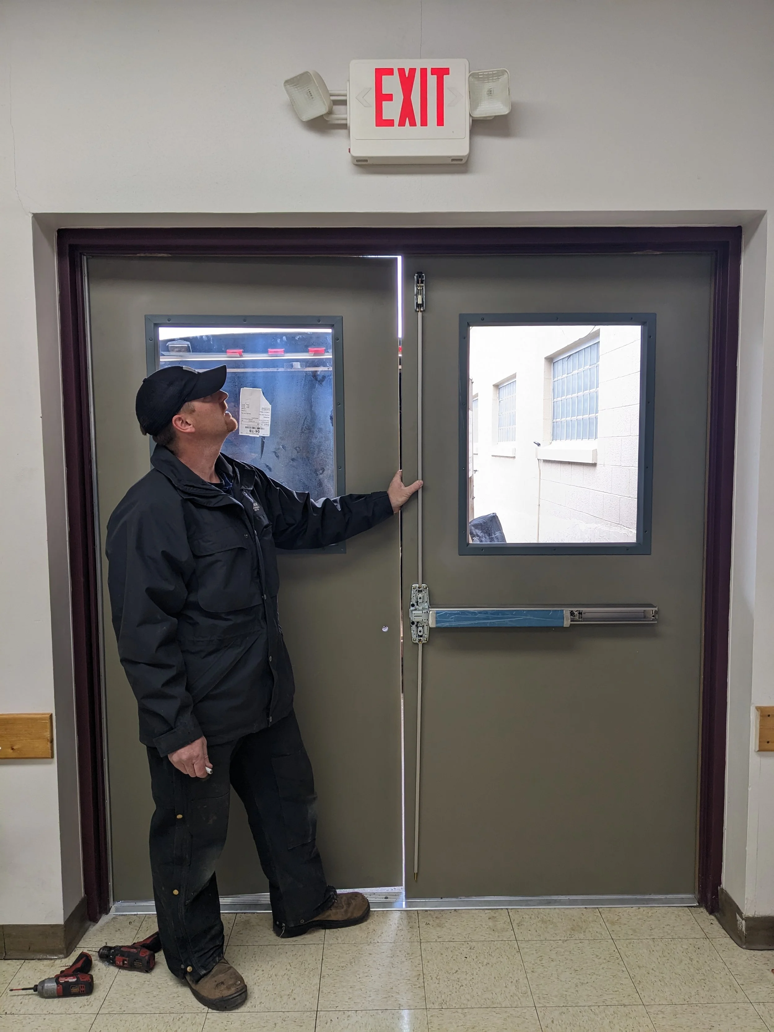 A person in black work clothes and a cap pushes a pair of gray emergency exit doors that have windows. There is a red and white "EXIT" sign above the doors and two lights on either side of the sign.