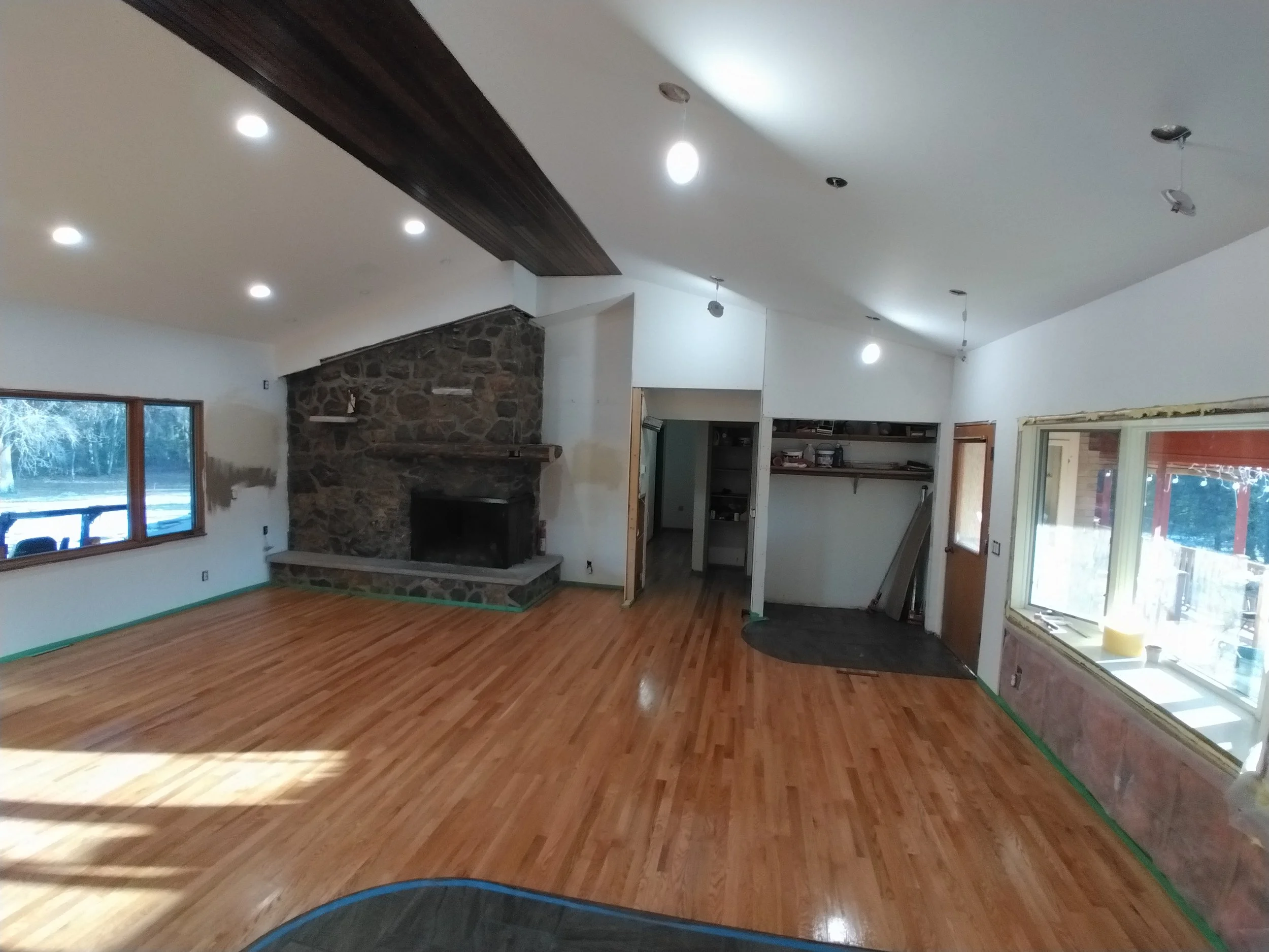 Living room undergoing renovation with hardwood floors, large windows, a stone fireplace, and ceiling lights. Some areas are still under construction or waiting for finishing touches.
