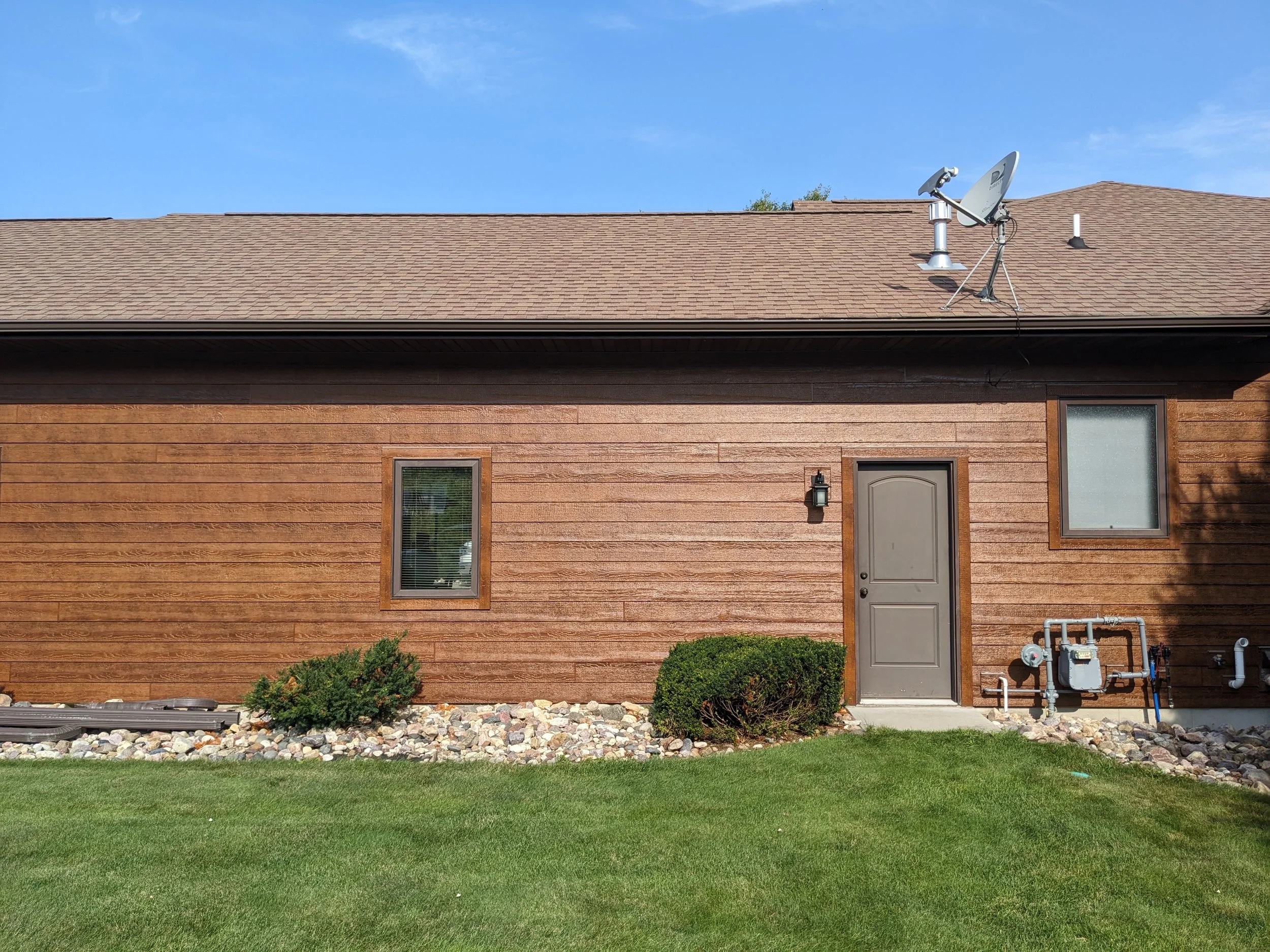 Rear exterior of a house with brown wooden siding, two windows, a gray door, and a satellite dish on the roof. There are small bushes and rocks in front of the house with green grass in the foreground.